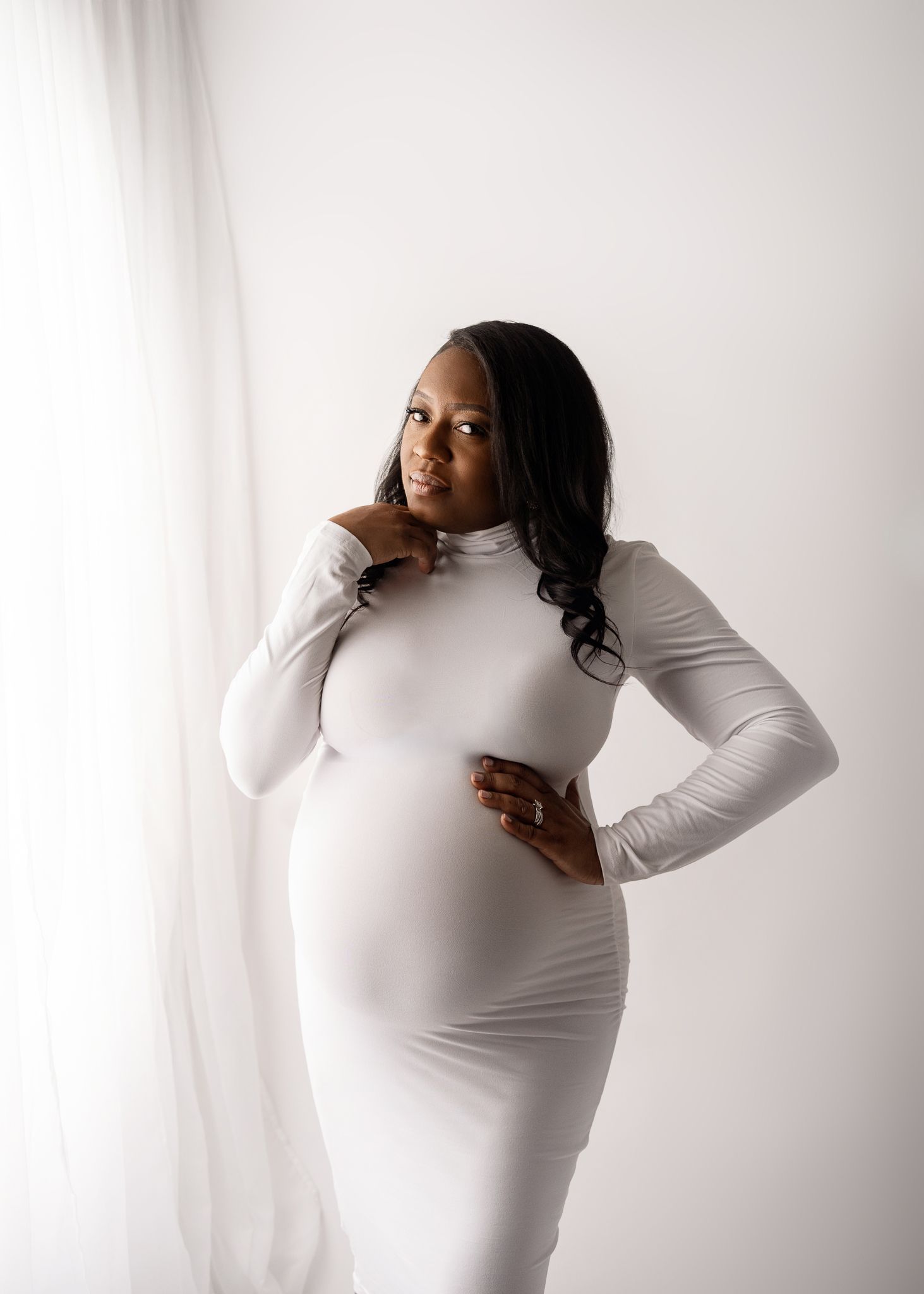 A pregnant person in a fitted white long-sleeve dress poses against a light background during a maternity photoshoot.