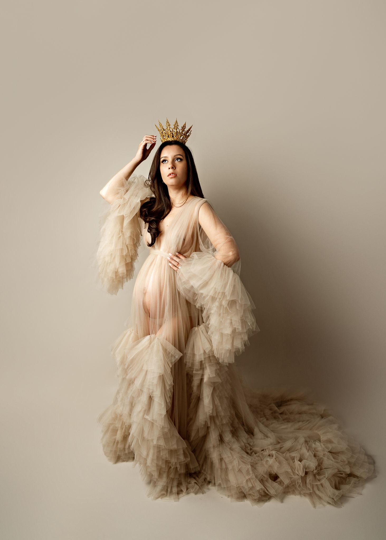 Ethereal maternity portrait featuring a flowing champagne-colored ruffled gown and delicate gold crown against neutral backdrop.