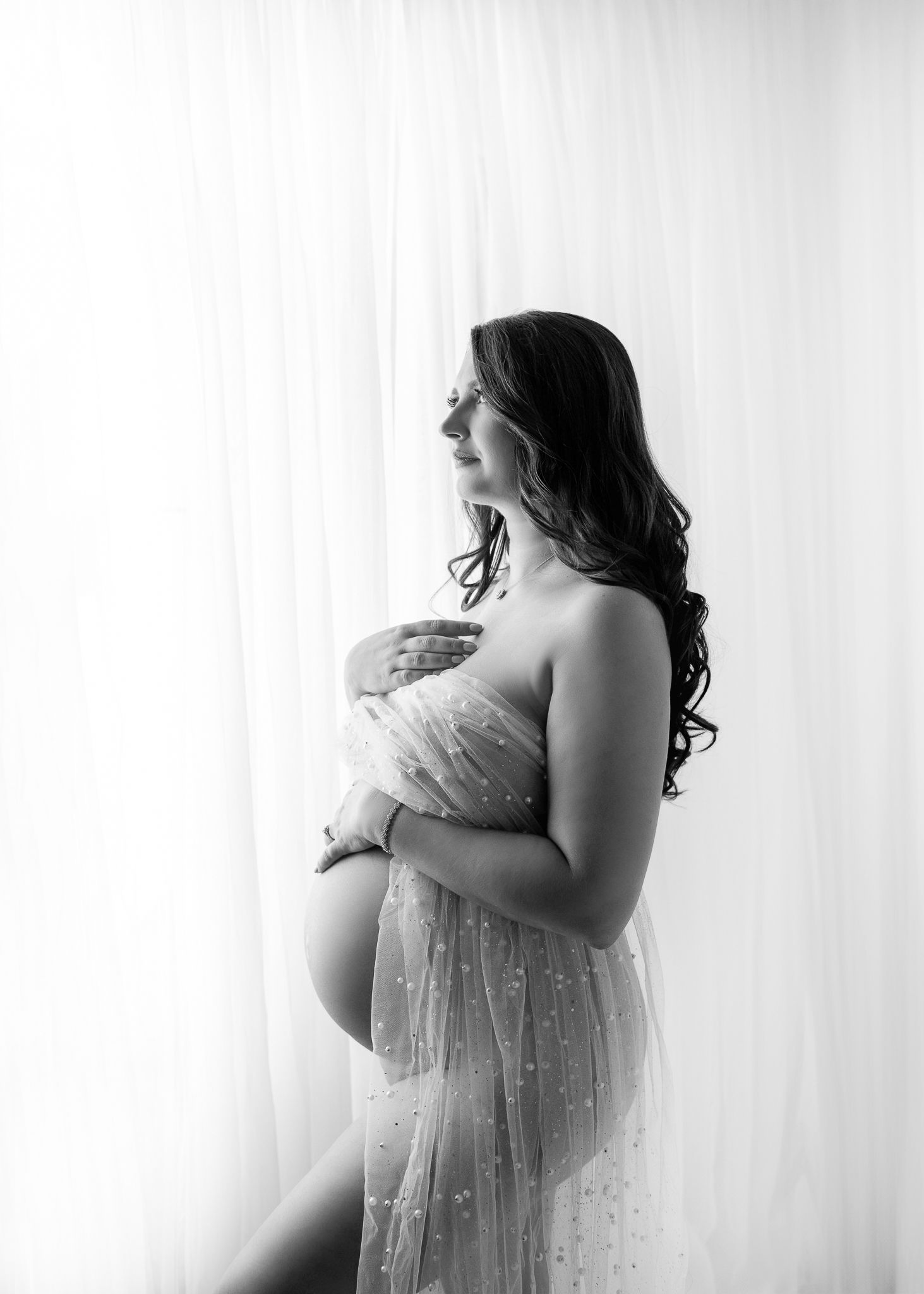 Black and white maternity portrait showing an expectant mother in a lace strapless gown against a light background.