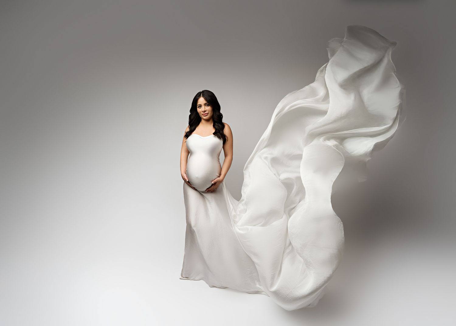 Flowing white fabric dramatically billows around posed figure in maternity photoshoot against gray backdrop.