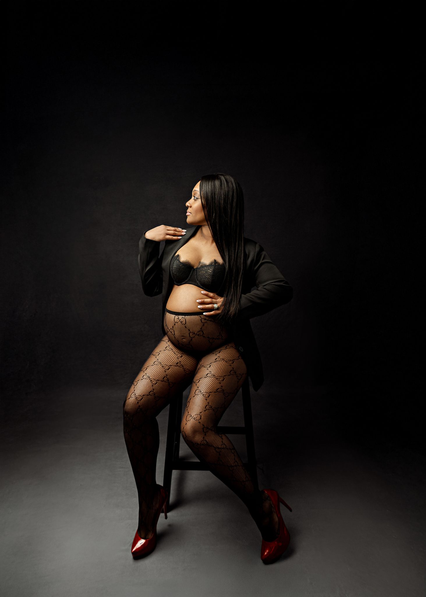 Artistic maternity portrait of figure in lace bodysuit and blazer posing on chair against dark background with dramatic lighting.