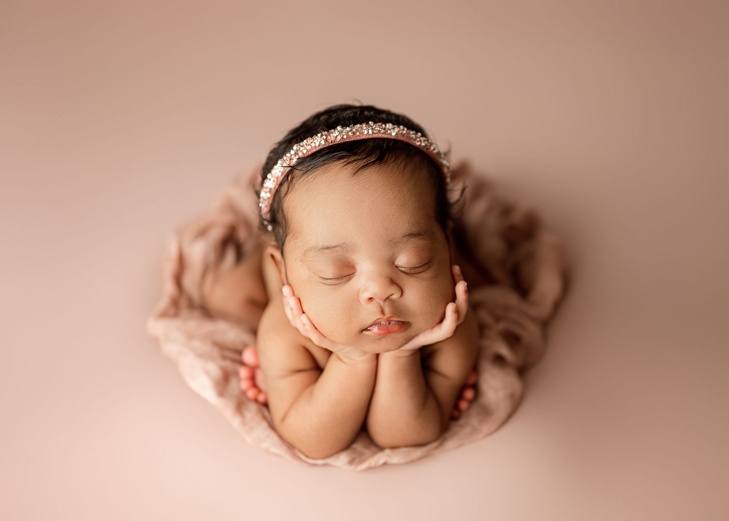 Peaceful sleeping newborn baby wearing delicate headband poses with hands under chin on pink blanket.
