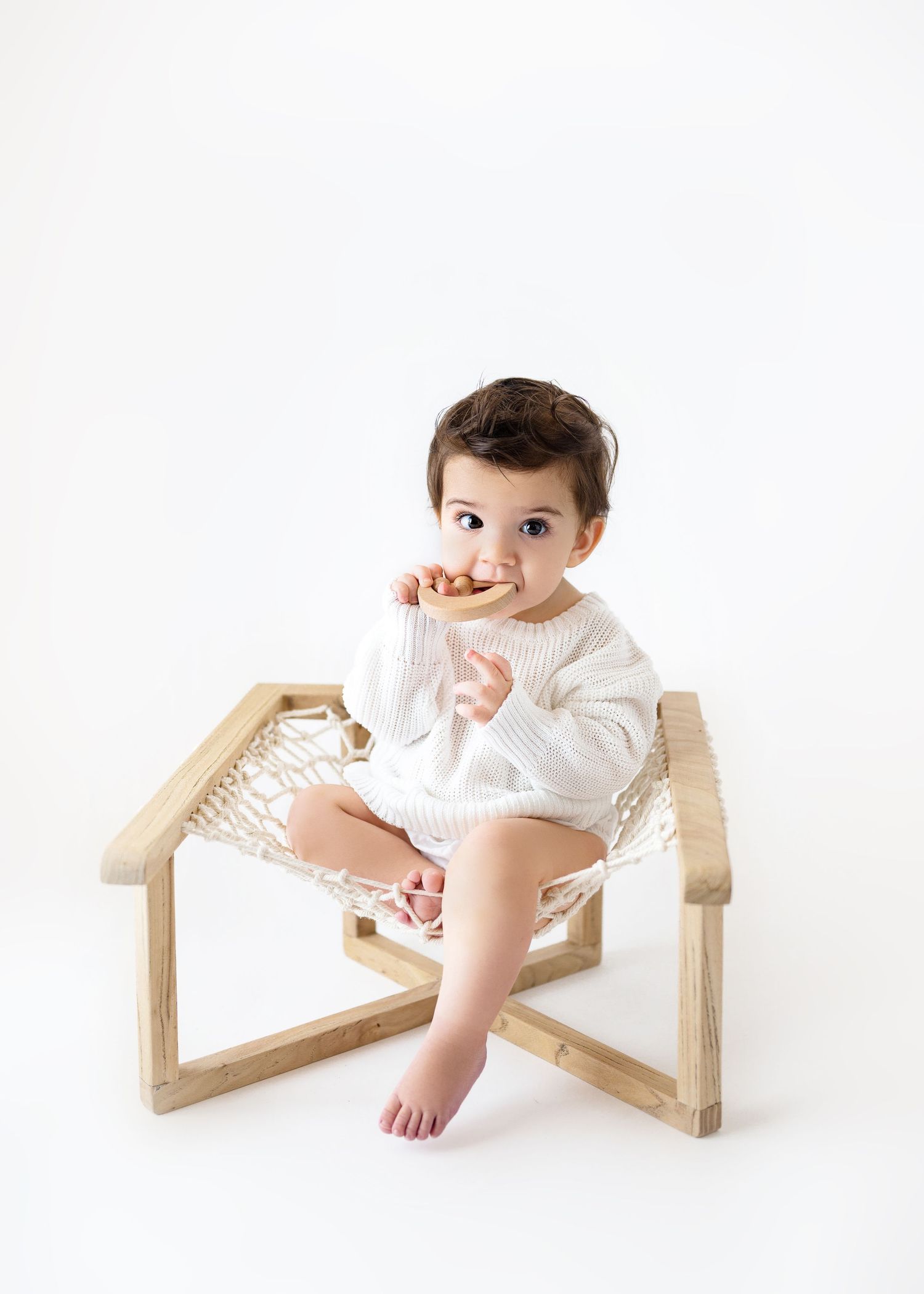 A baby wearing a white sweater sits in a modern wooden chair with woven macrame details on a white background.