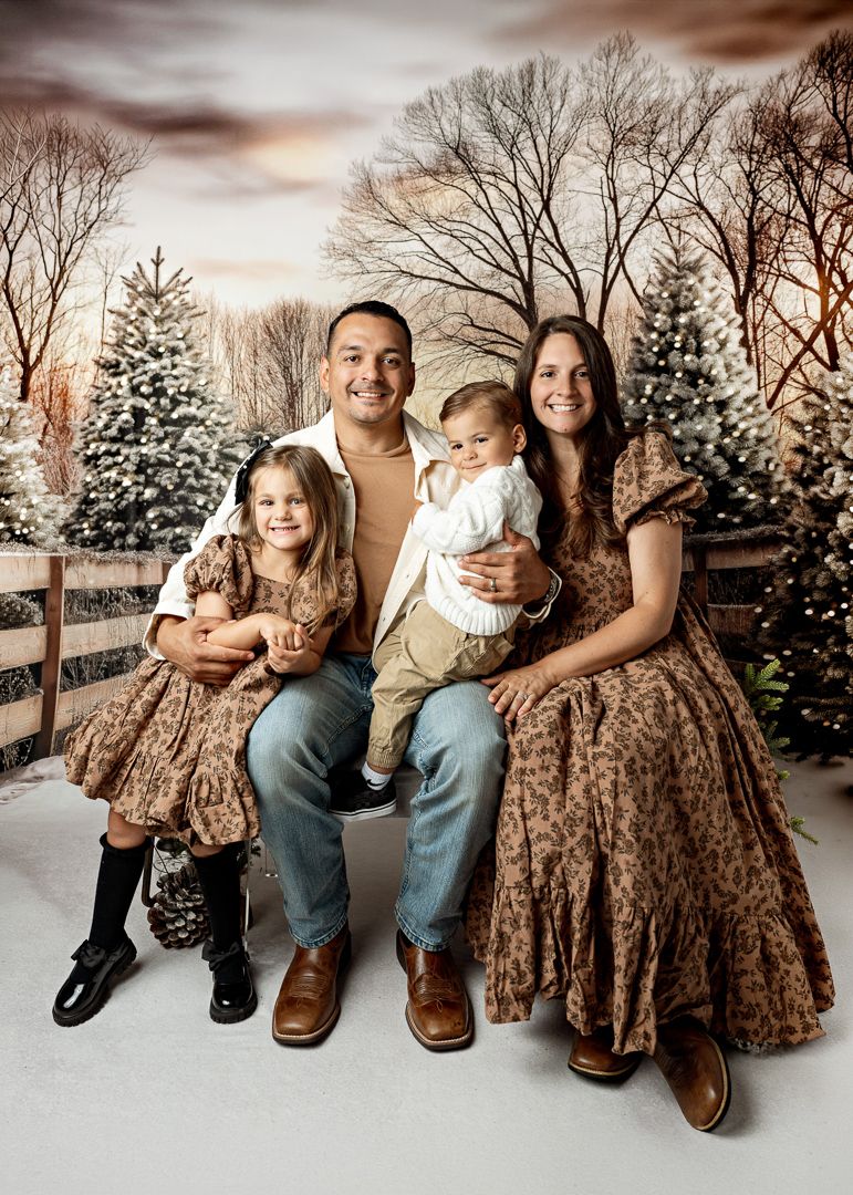 Family of four poses together in matching brown outfits against a snowy Christmas tree backdrop.