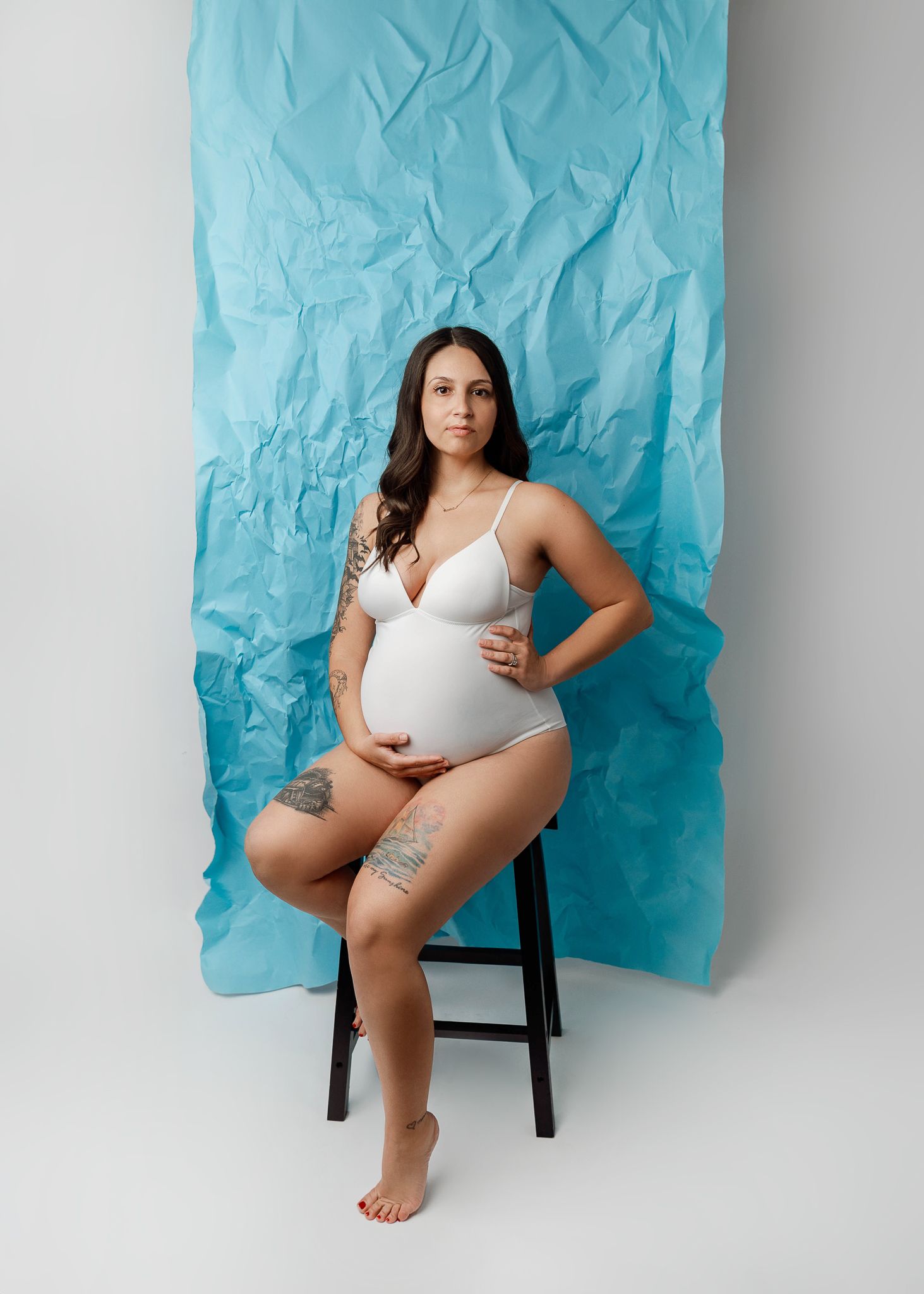 Maternity photo of woman in white bodysuit posing on stool against textured light blue backdrop.