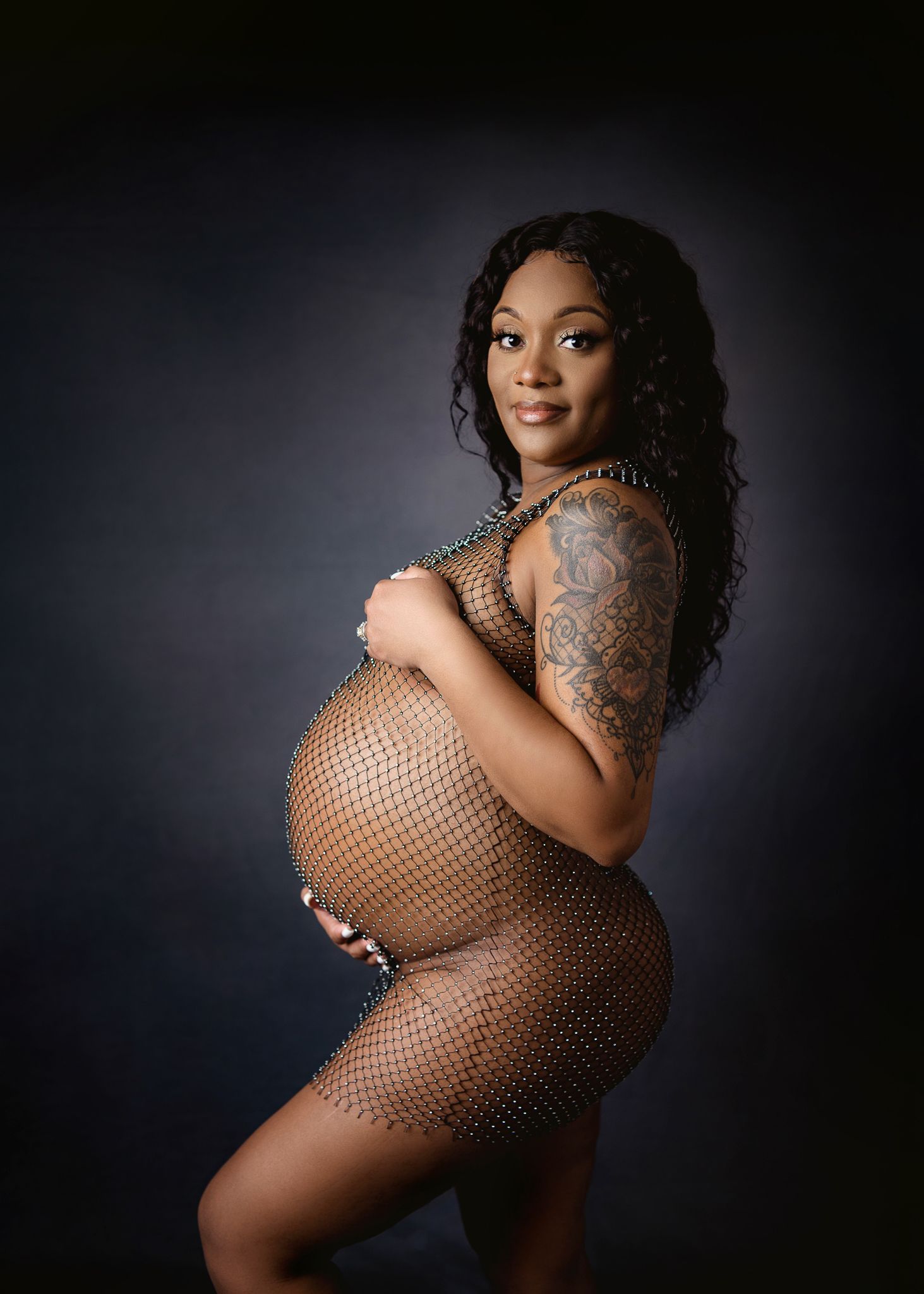 Artistic maternity photo featuring a form-fitting fishnet dress against a dark background.