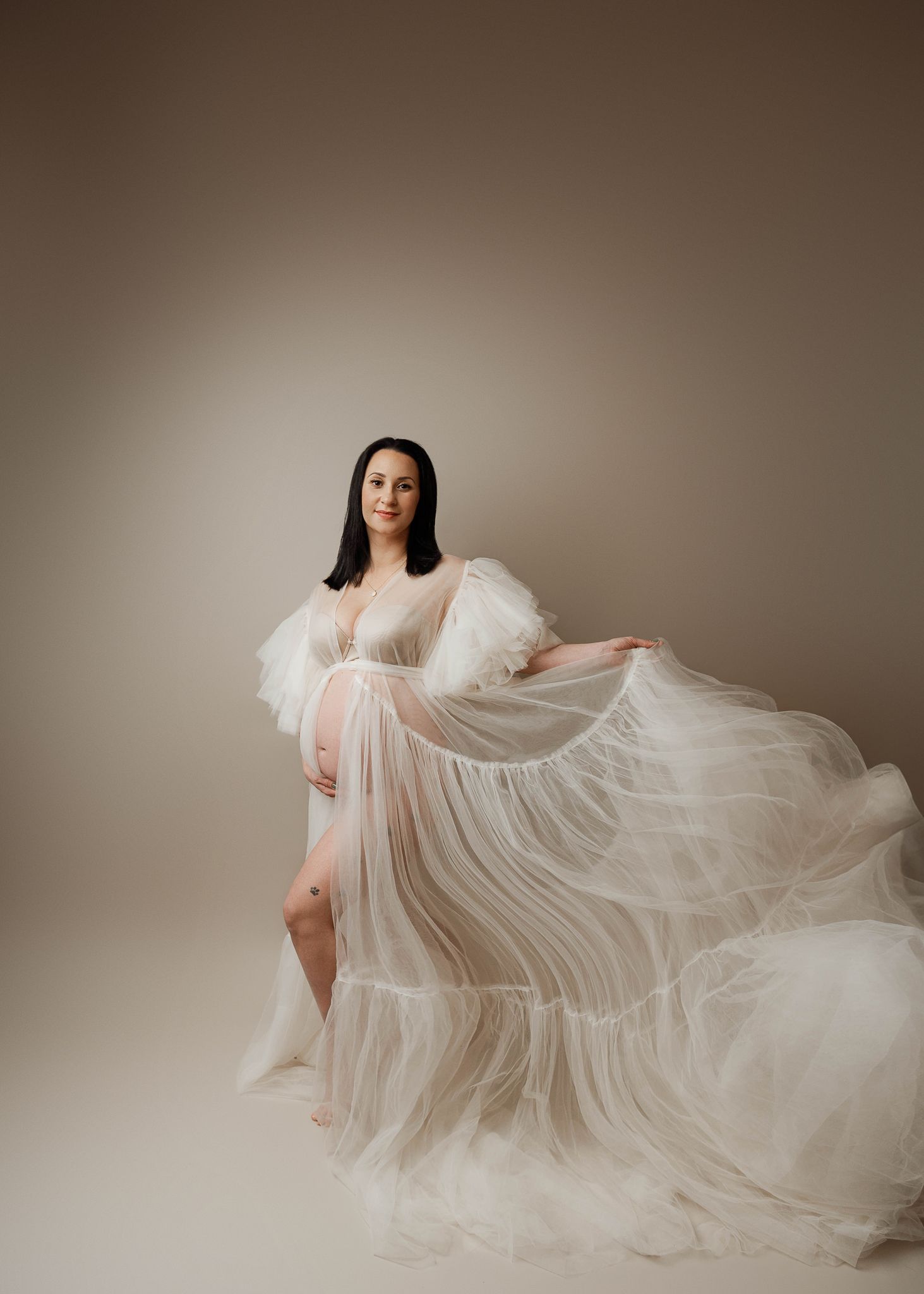 An ethereal maternity photo featuring a flowing sheer white gown with dramatic movement against a neutral backdrop.