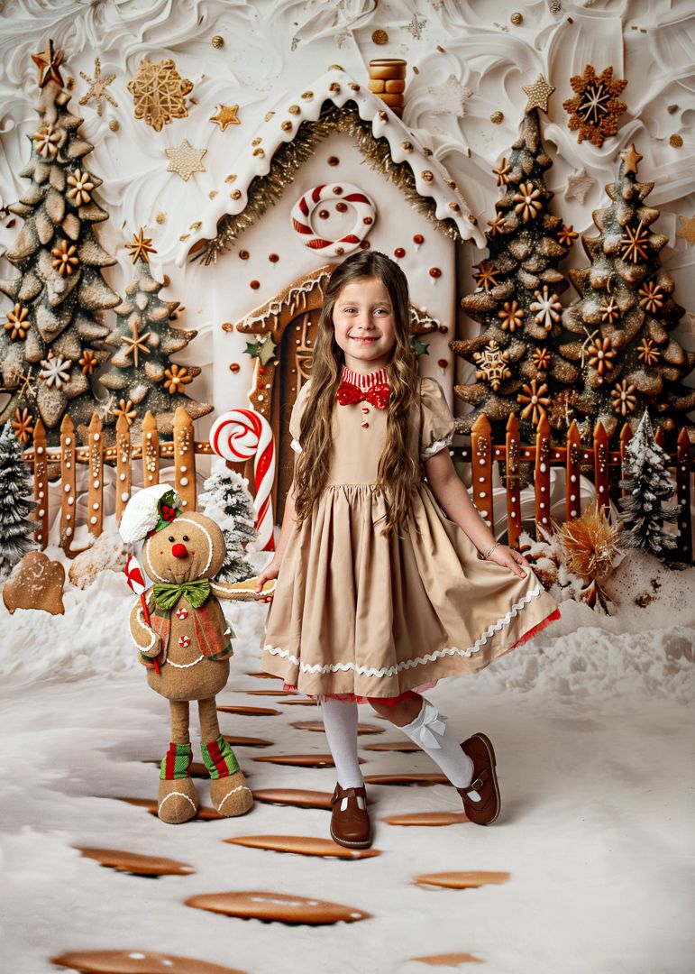 Young girl in gingerbread dress poses before festive holiday backdrop with candy cane house and gingerbread man plush toy.