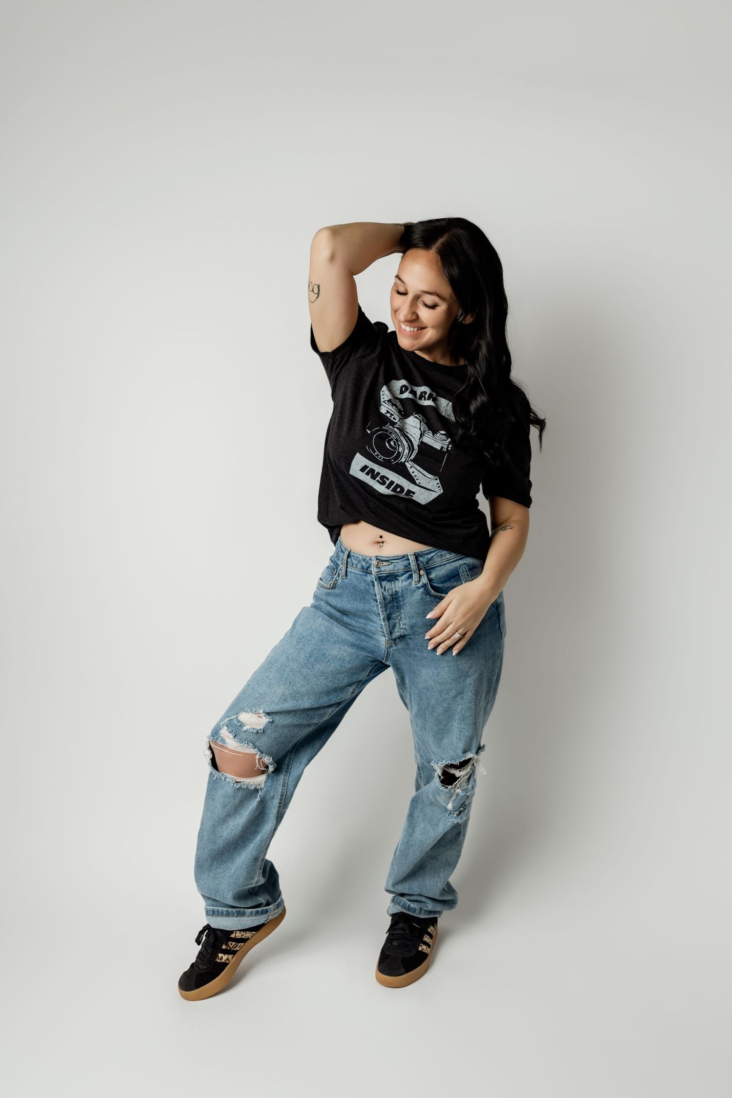 Woman in black graphic tee and ripped jeans posing dynamically with hand in dreadlocks on white background.