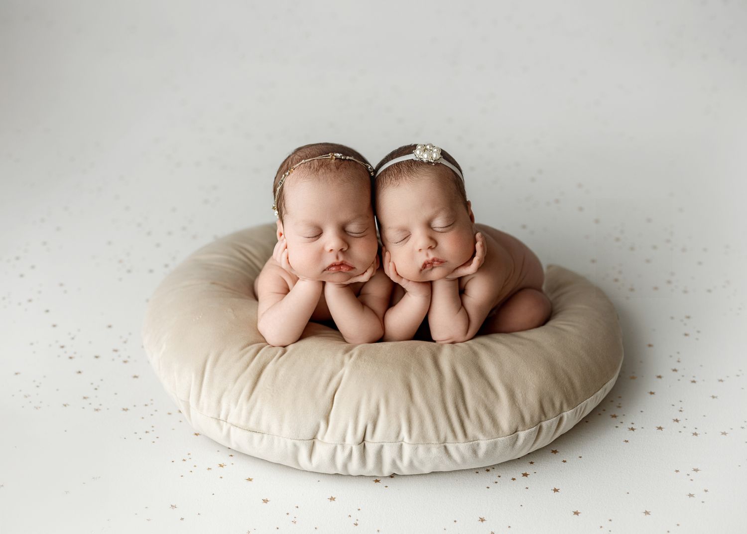 Two sleeping newborn babies posed together on a round cream-colored cushion in a professional studio setting.