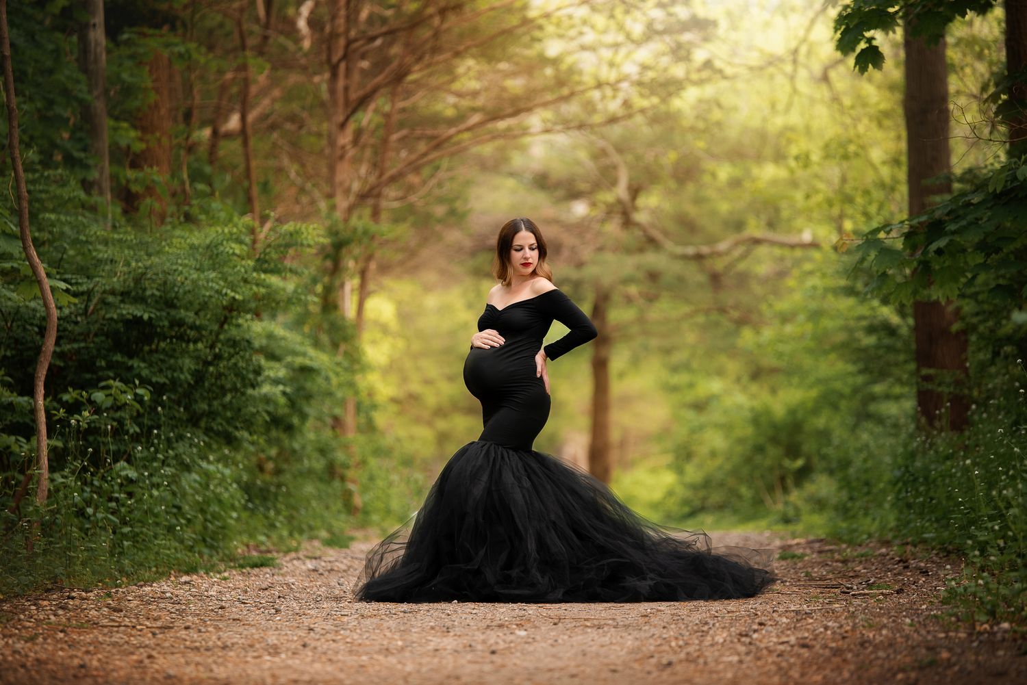 A flowing black maternity gown trails elegantly along a wooded forest path in magical golden sunlight.