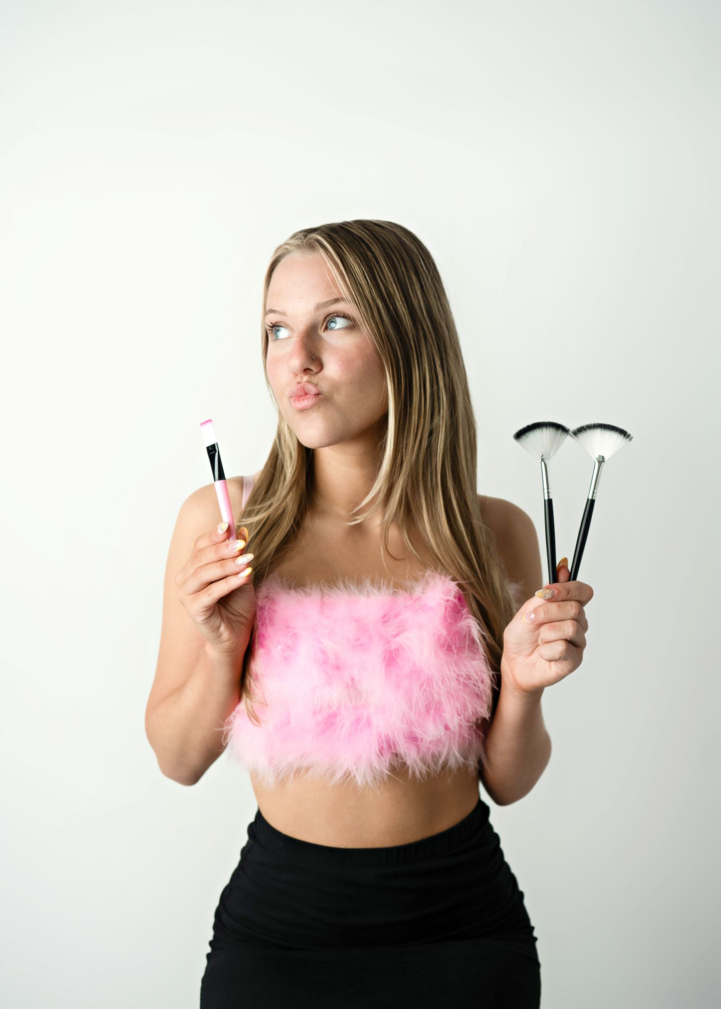 Woman in pink feathered crop top holding makeup brushes against white background.