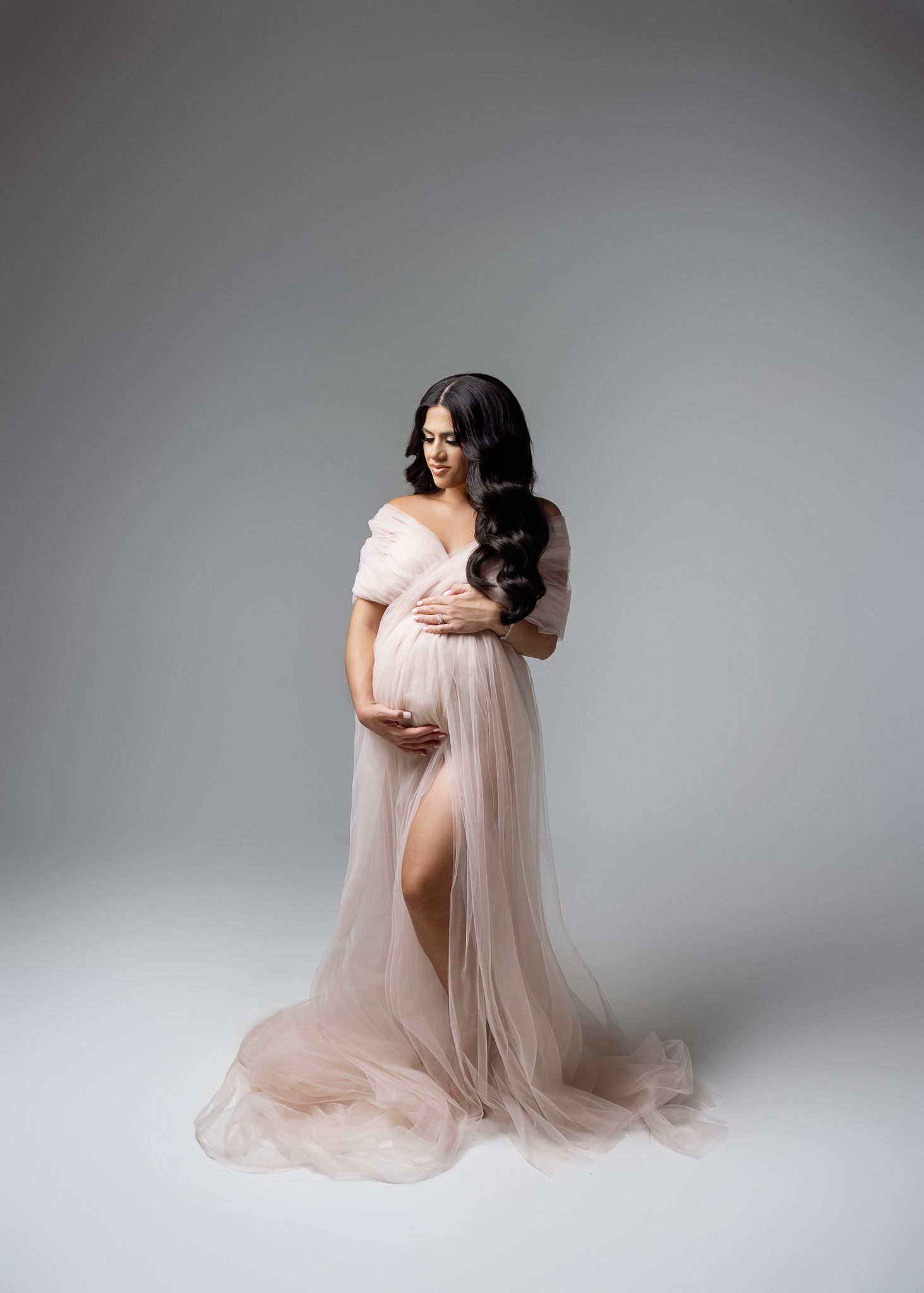 Elegant maternity photo featuring a flowy blush tulle gown with off-shoulder design against neutral grey background.
