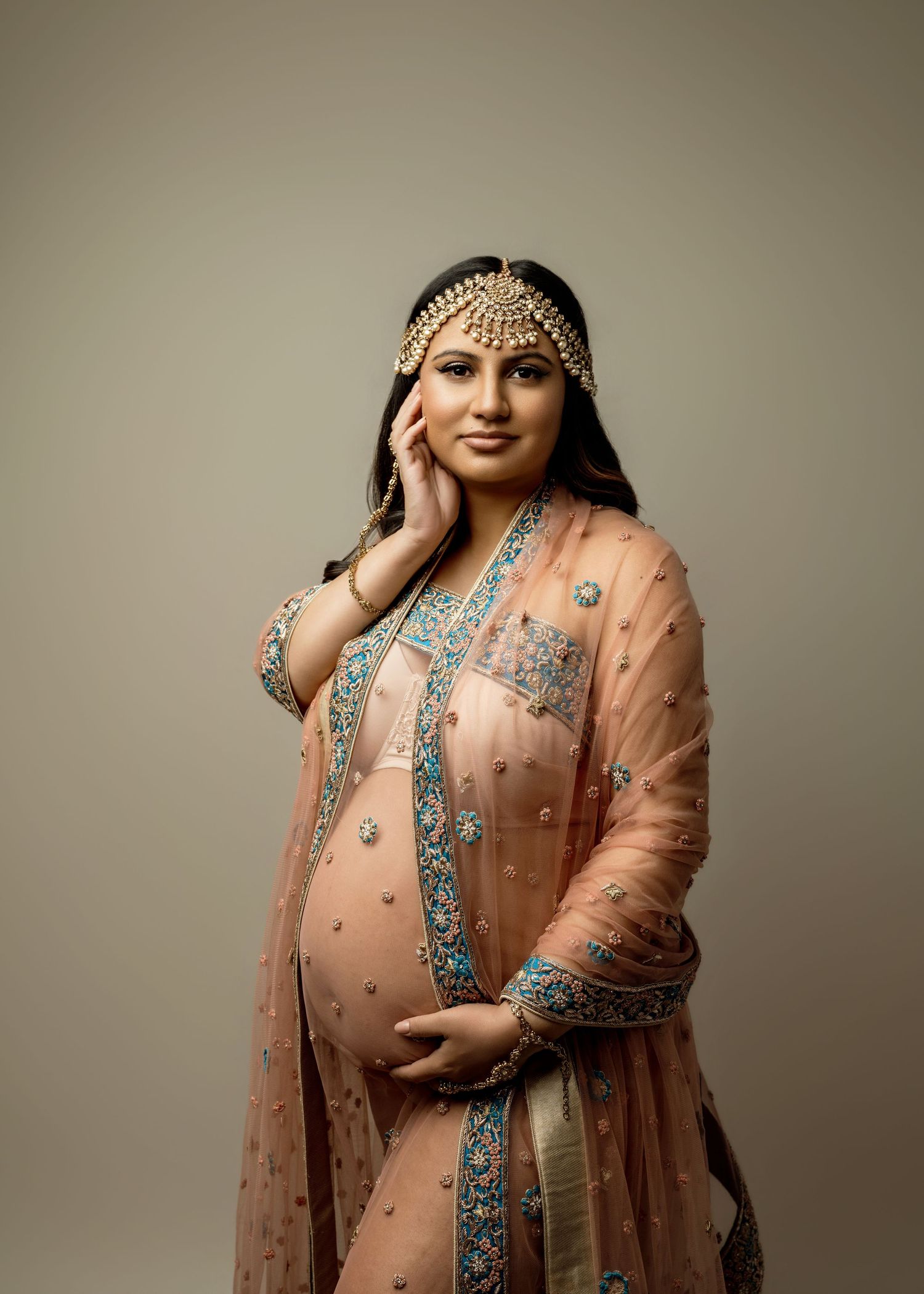 Pregnant woman in elegant peach embroidered Indian salwar kameez with gold maang tikka, cradling baby bump in studio portrait.