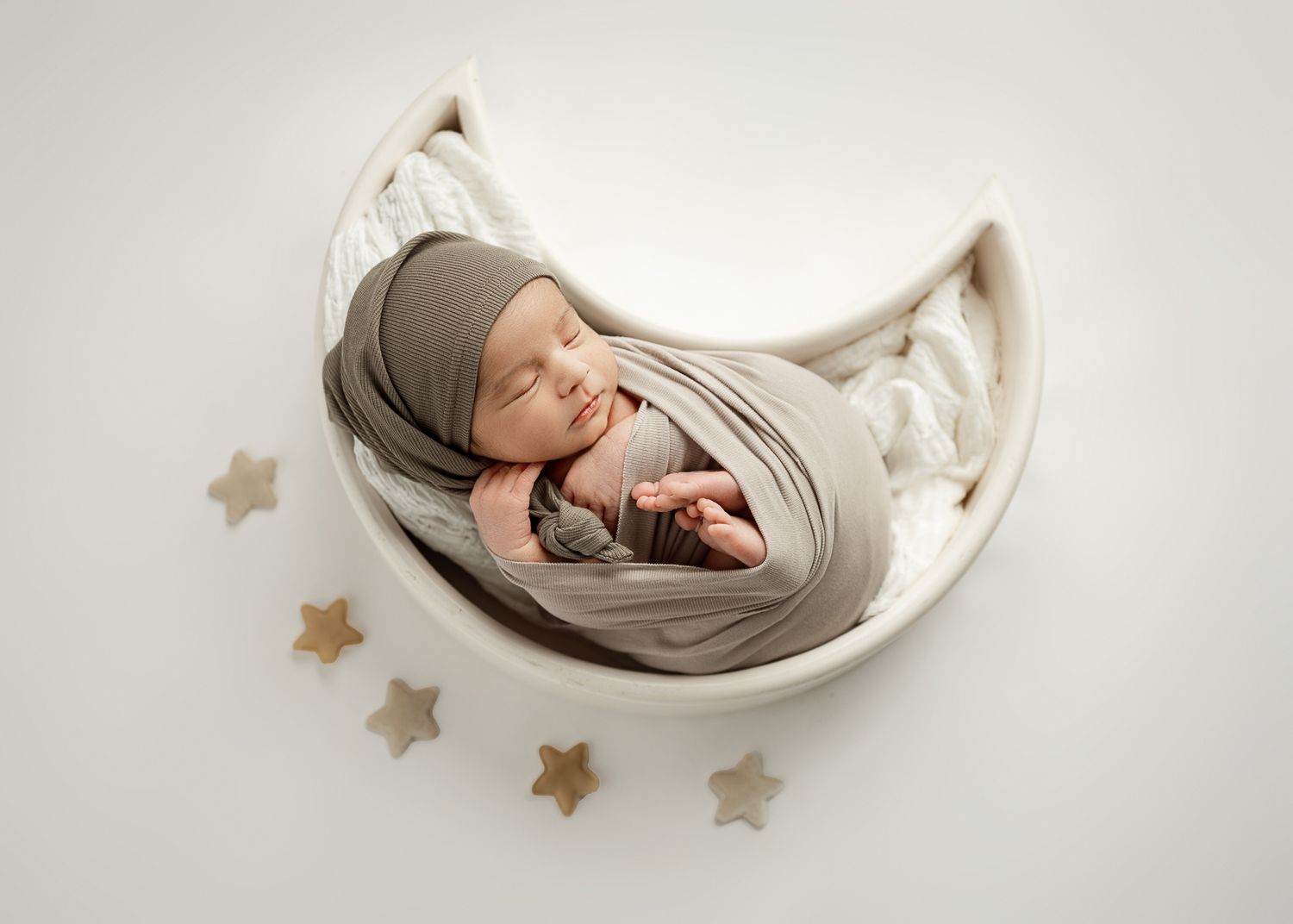 Peaceful sleeping newborn swaddled in soft gray fabric lying in curved white bowl with decorative gold stars scattered around.