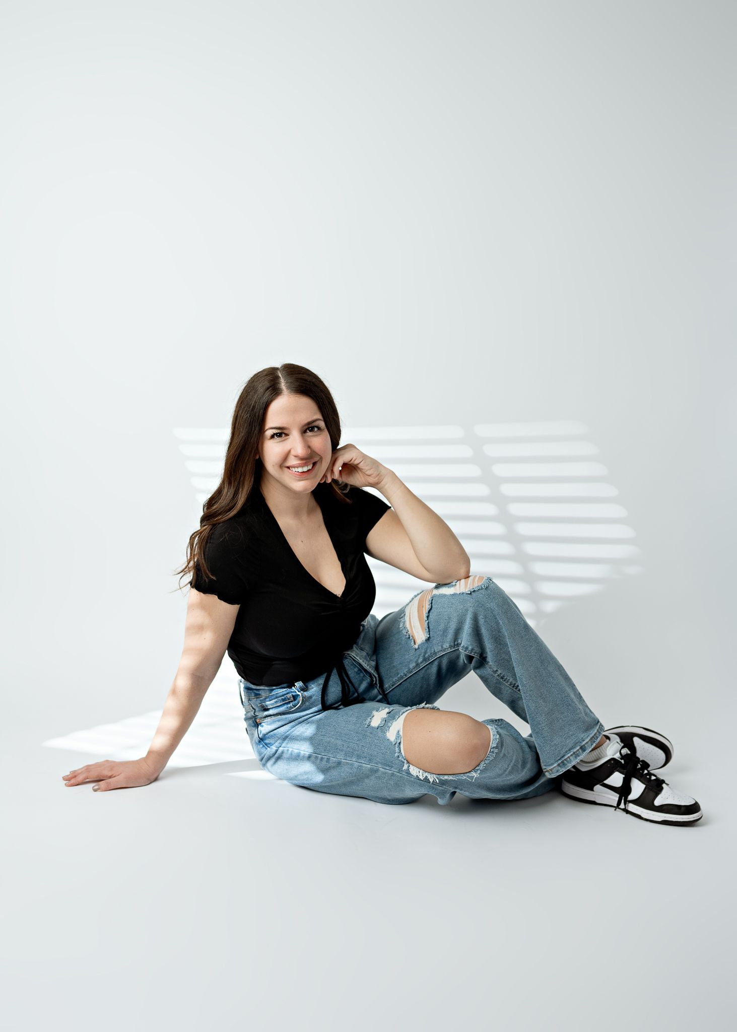 Person in casual black top and distressed jeans sits on white floor with window light casting shadows.
