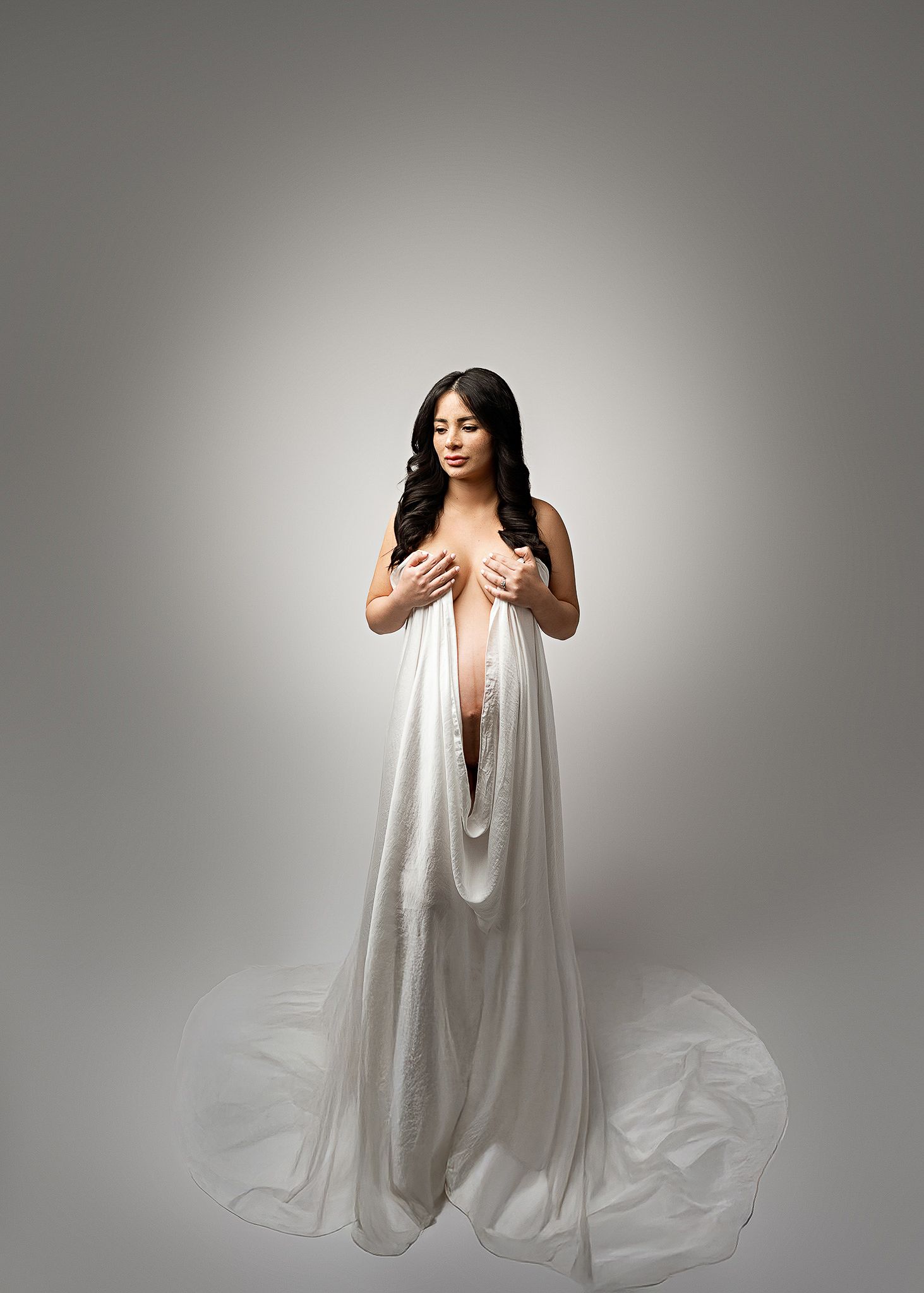 Figure draped in flowing white fabric stands gracefully against neutral grey background in artistic portrait study.
