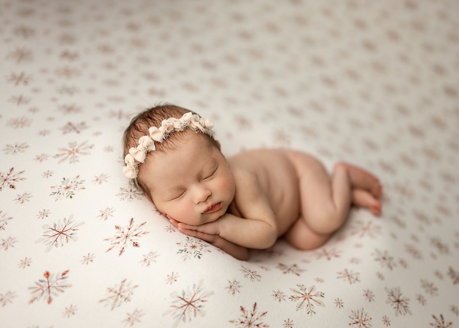 Sleeping newborn baby wearing delicate floral headband rests peacefully on patterned blanket during professional photo session.