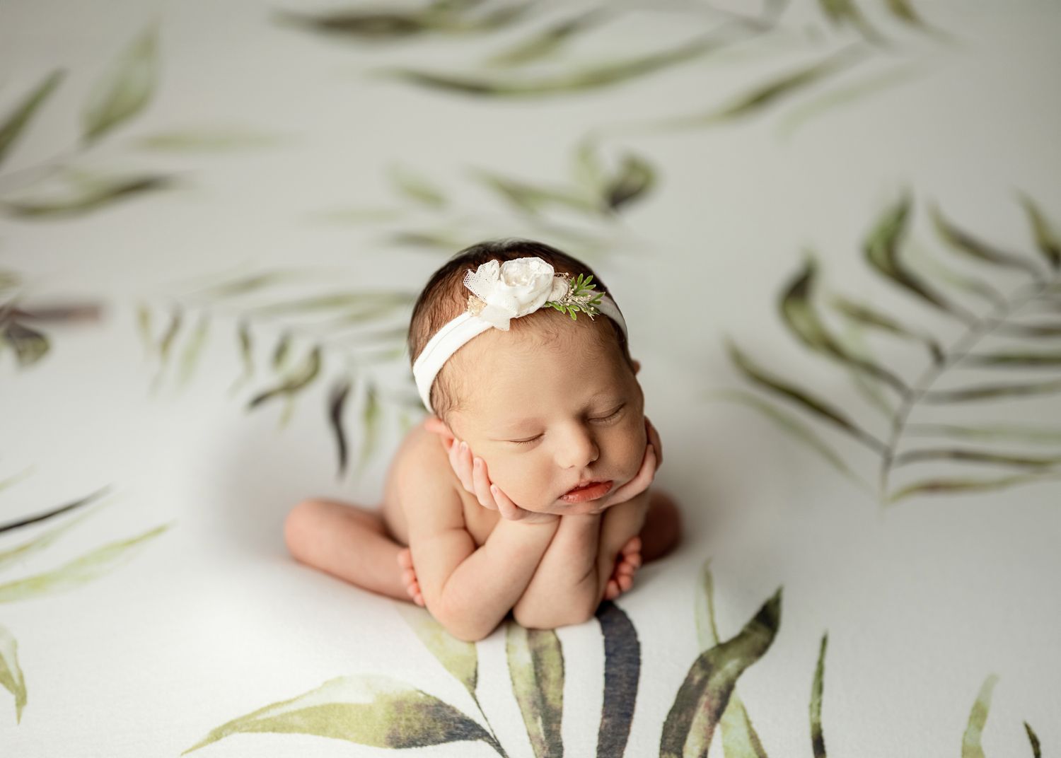 A sleeping newborn baby in white swaddle lies on a green floral patterned background in soft natural light.