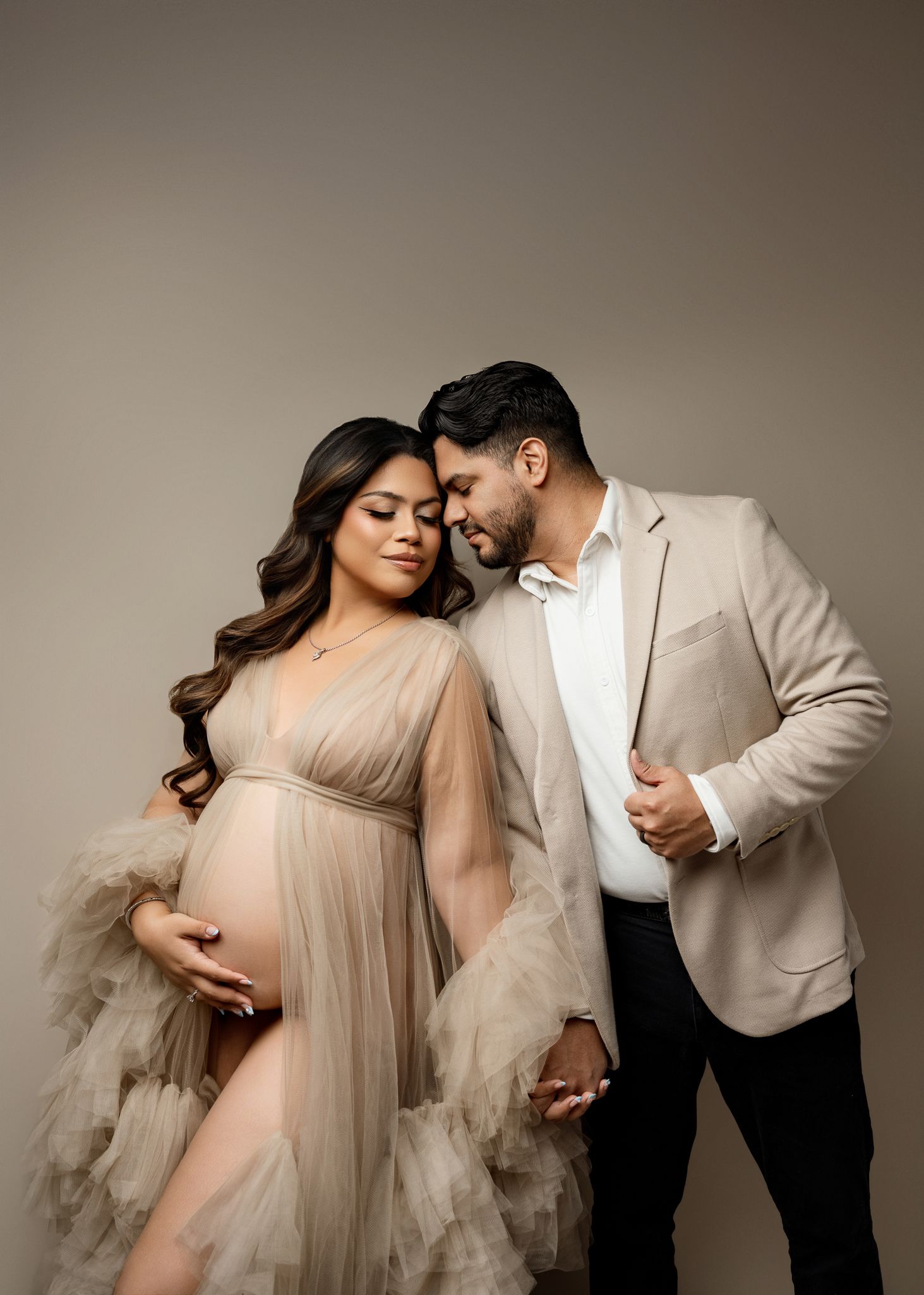Romantic maternity photoshoot featuring couple in neutral tones with flowing tulle dress against beige backdrop.