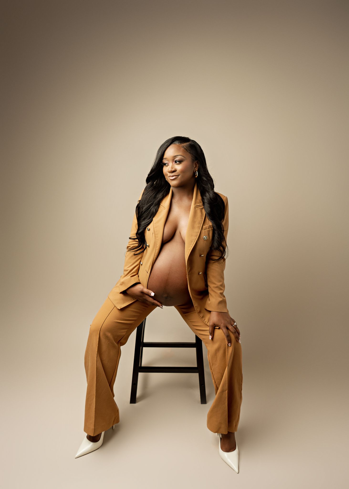 A stylish maternity portrait featuring a brown pantsuit and white heels against a neutral beige backdrop.