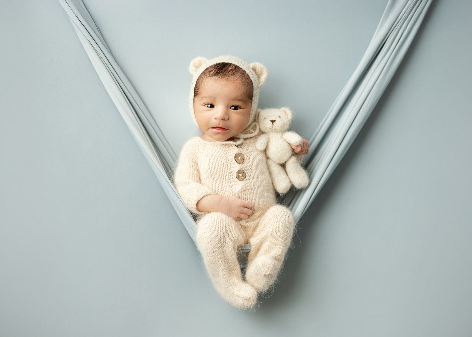 Baby wearing cream onesie and bear ear hat lies in fabric hammock with matching teddy bear against gray backdrop.
