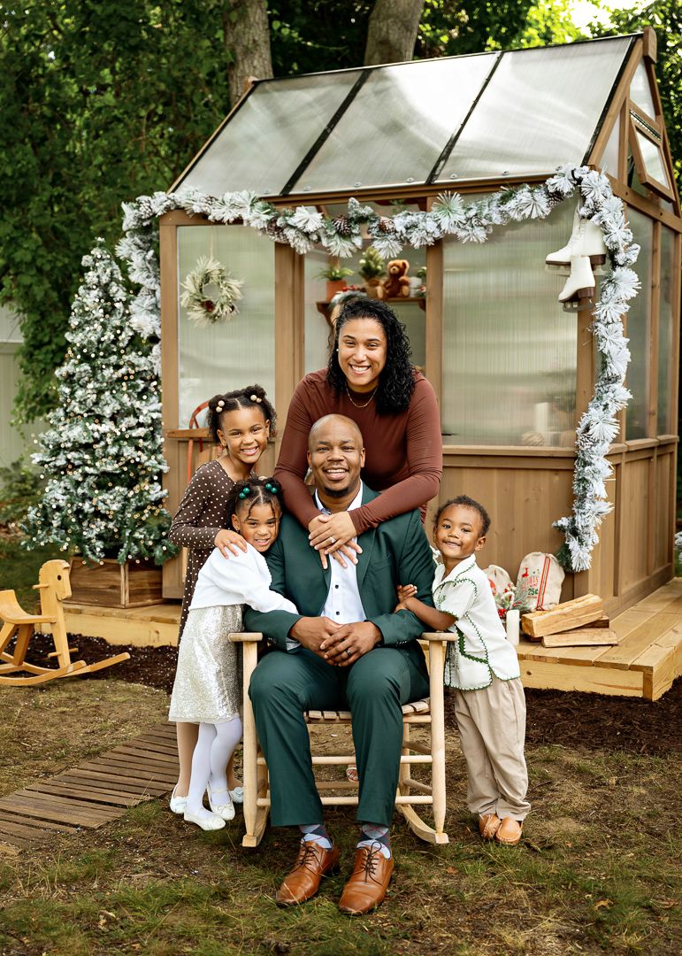 A joyful family of five poses in front of a decorated greenhouse with Christmas garland and a festive tree.