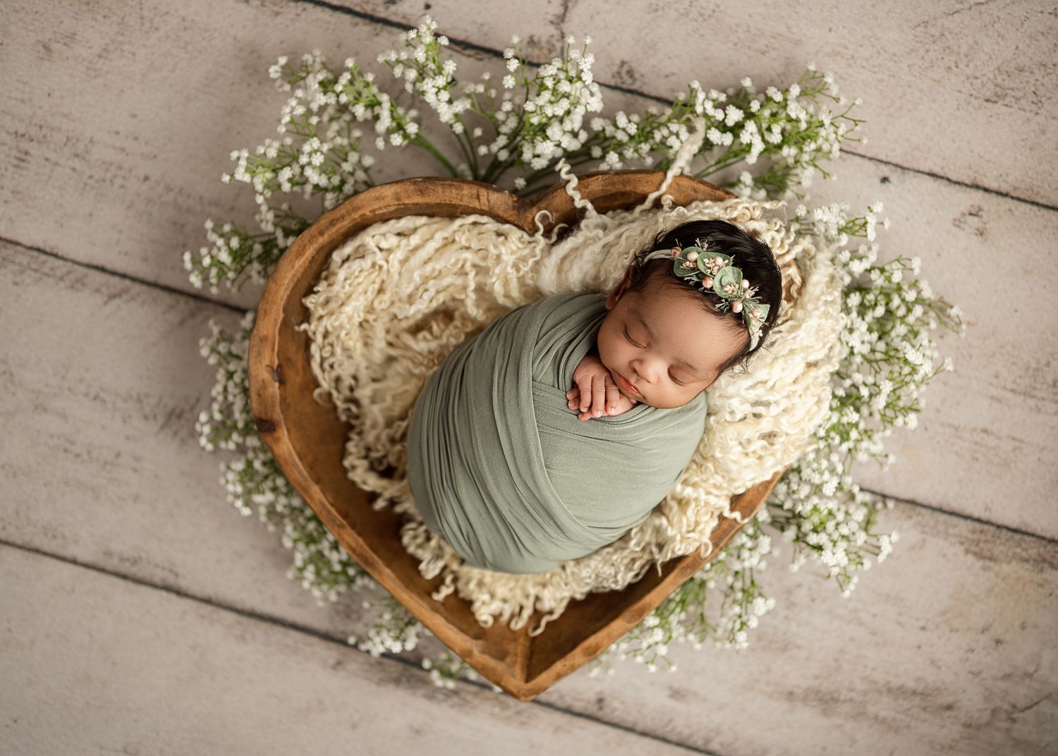 Newborn wrapped in mint green swaddle sleeps peacefully in heart-shaped basket surrounded by delicate baby's breath flowers.