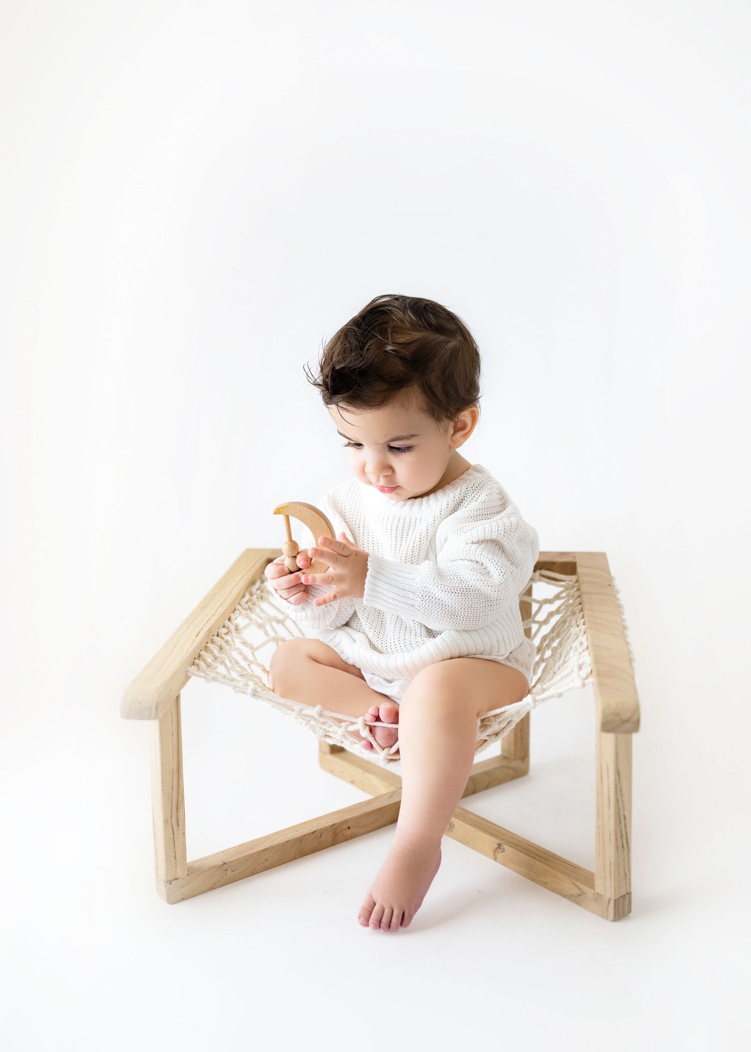 Small child in white romper sits on modern wooden frame chair with woven seat against white background.