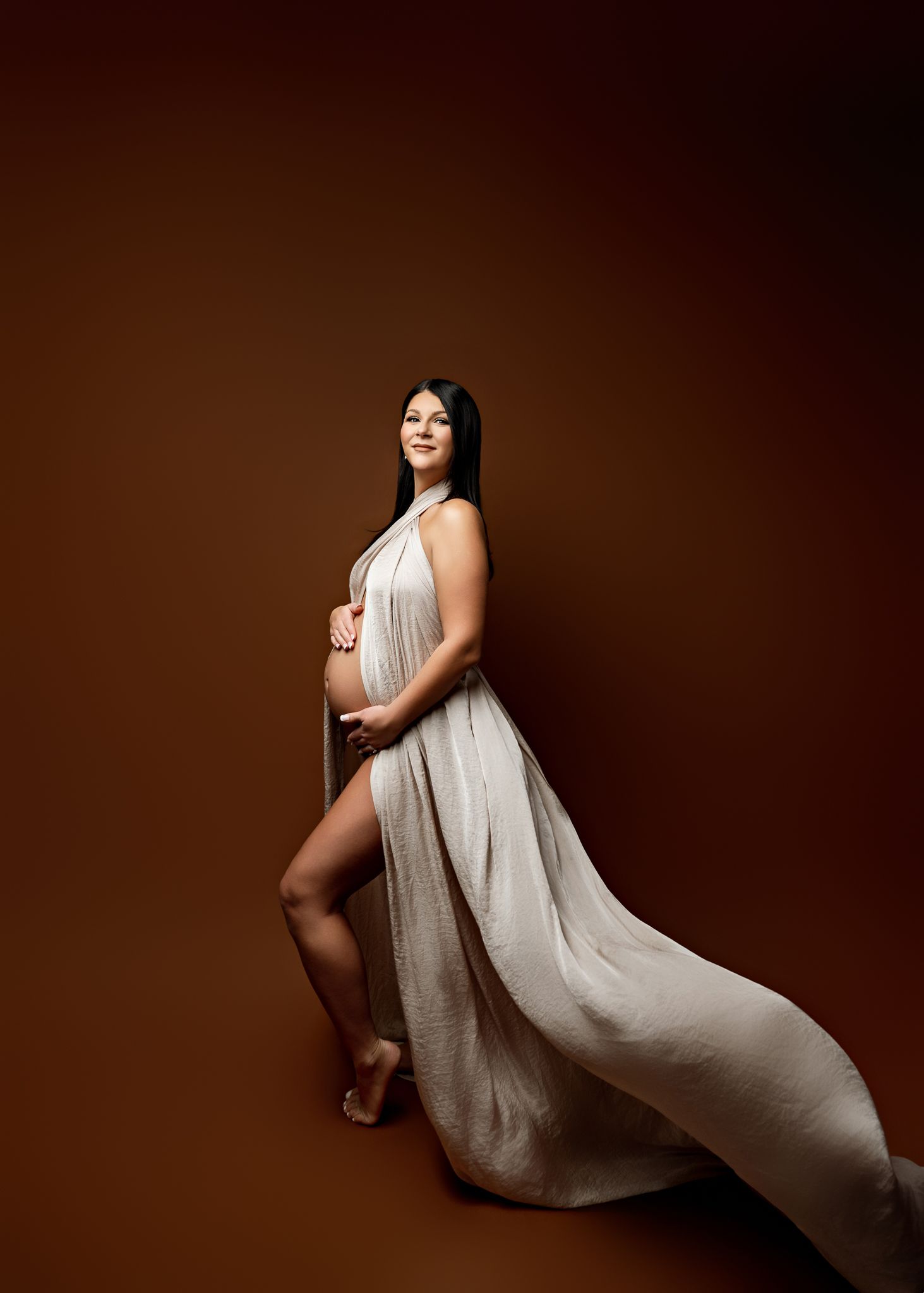 Artistic maternity portrait featuring a flowing beige dress against a dark brown studio backdrop.