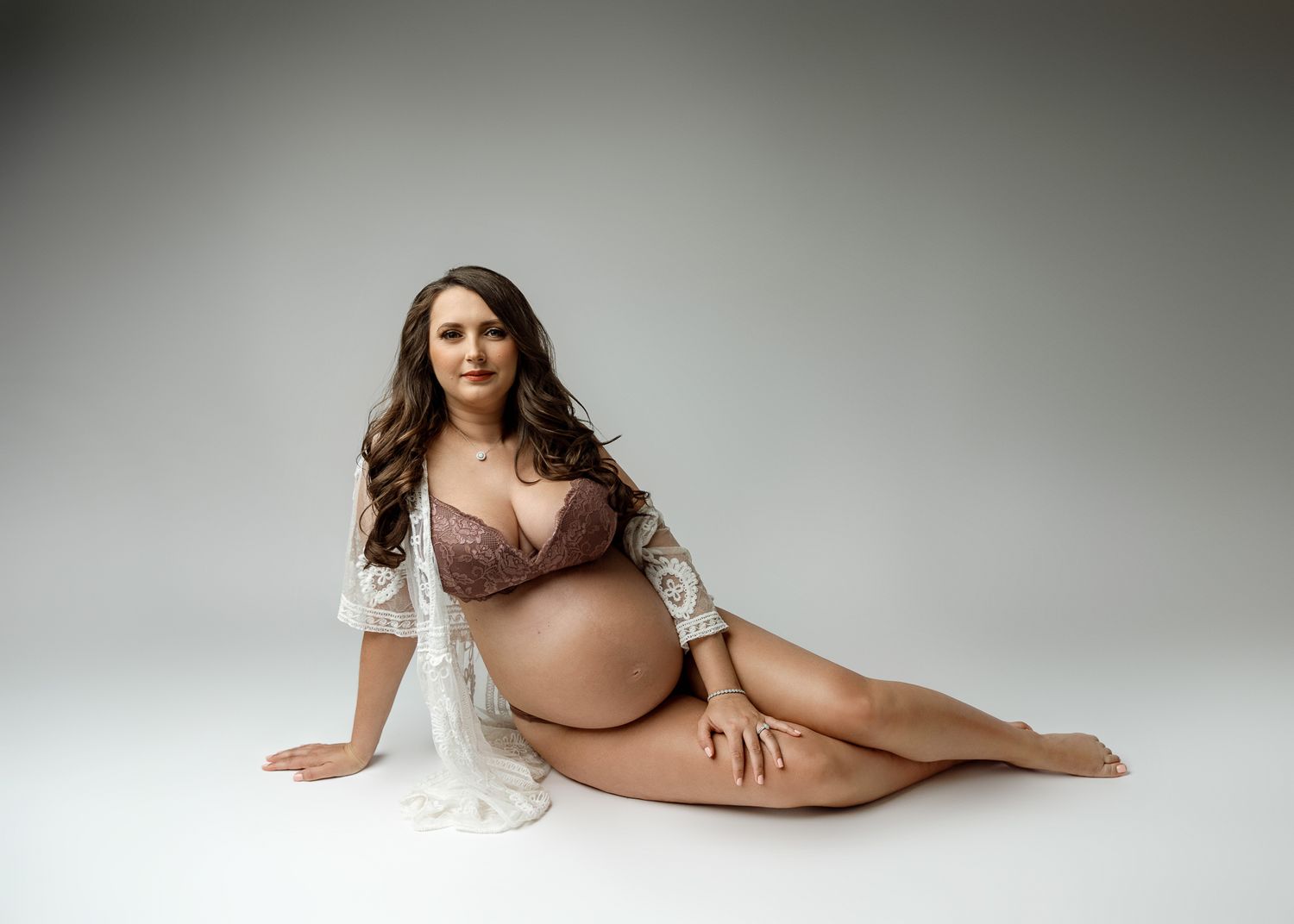 Maternity portrait of expectant mother in lingerie and lace robe posing on studio floor.