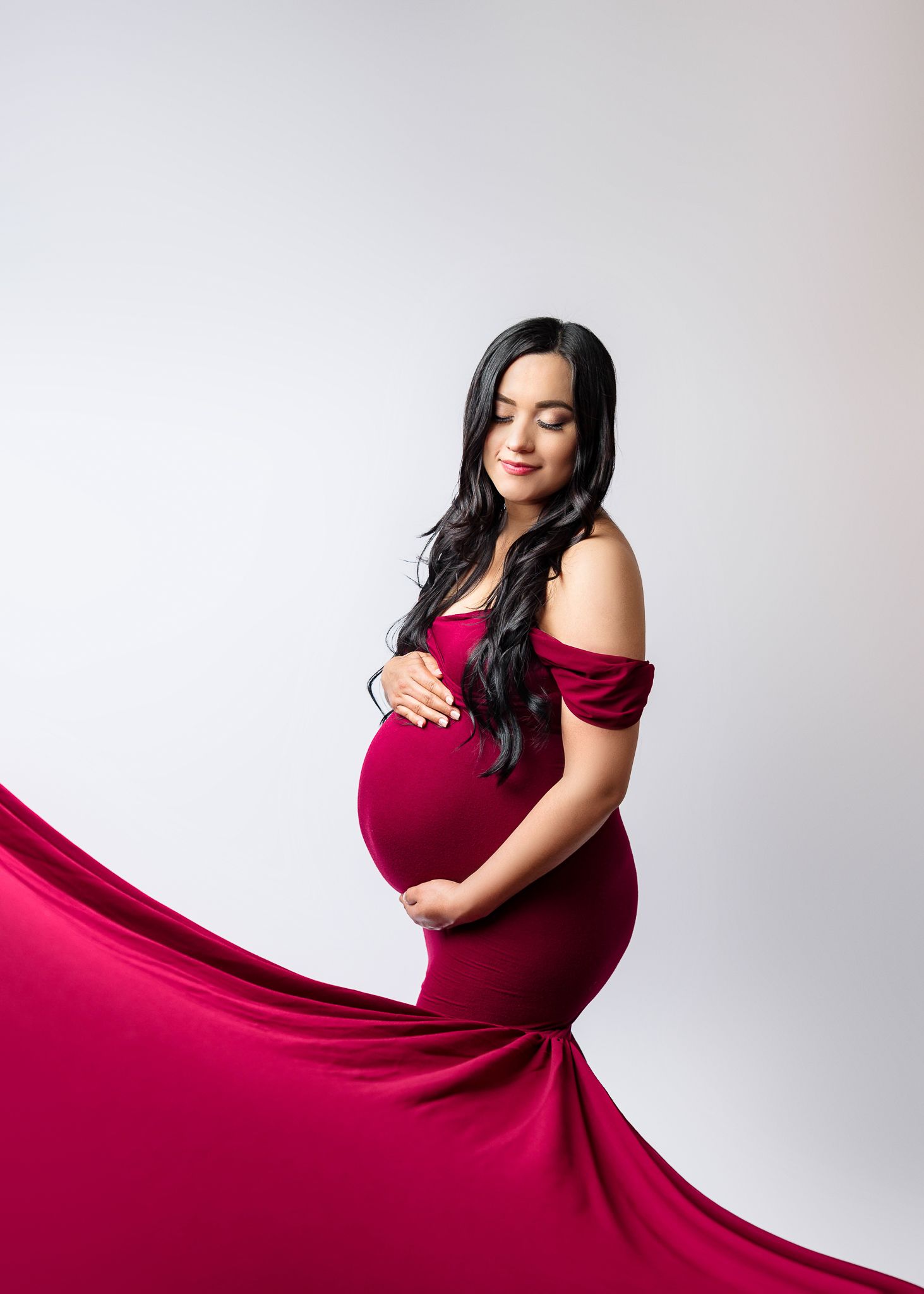 Elegant maternity photo of an expectant mother in a flowing red off-shoulder gown against a white background.