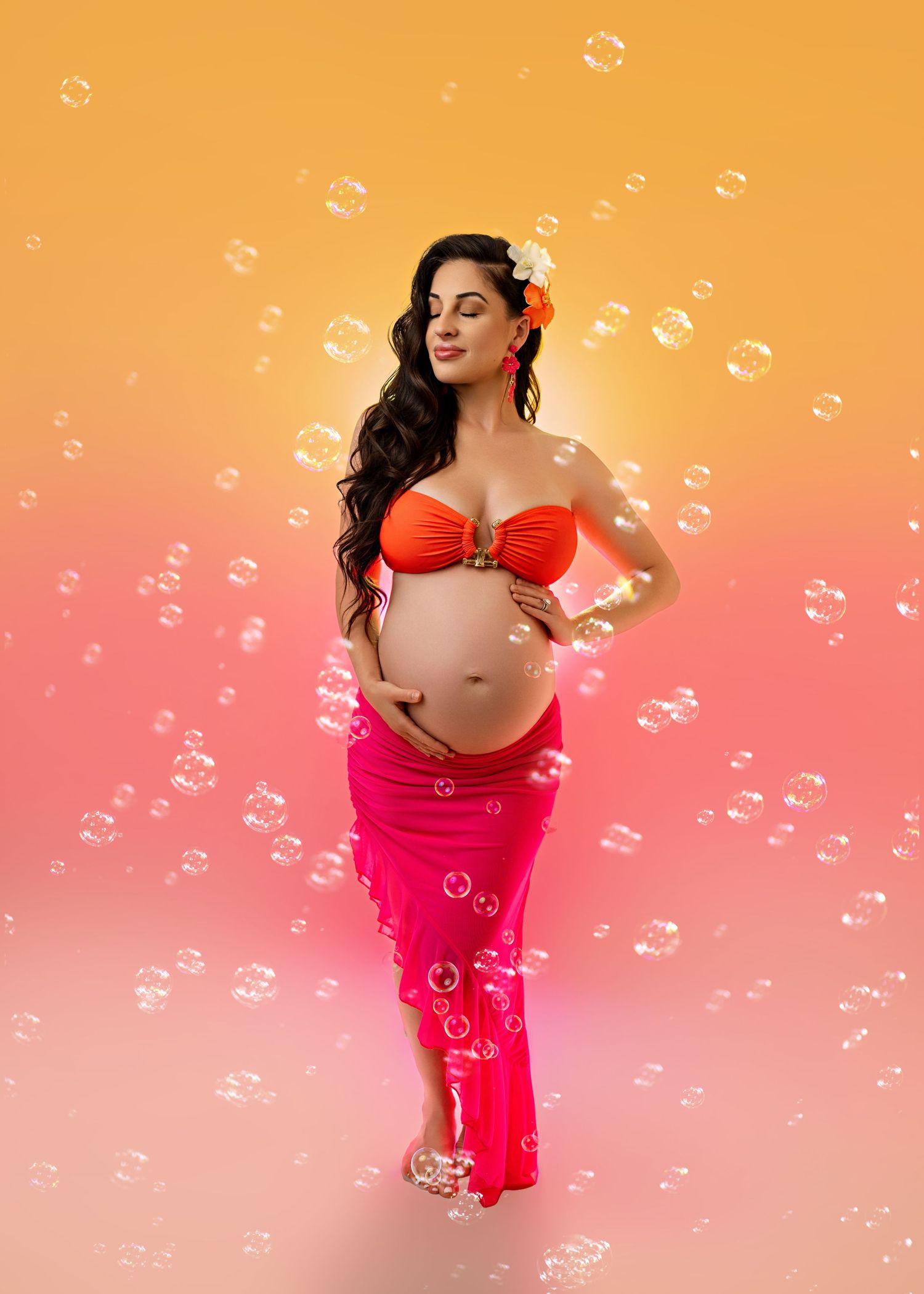Pregnant woman in red bikini top and pink skirt poses gracefully surrounded by floating bubbles on gradient background.