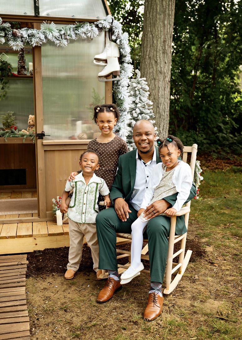 A smiling father in a green suit sits in a rocking chair with three young children during a festive Christmas outdoor photo.