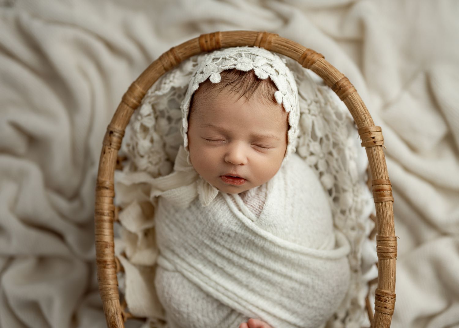 Newborn baby peacefully sleeping in a wooden basket wrapped in soft white fabric.