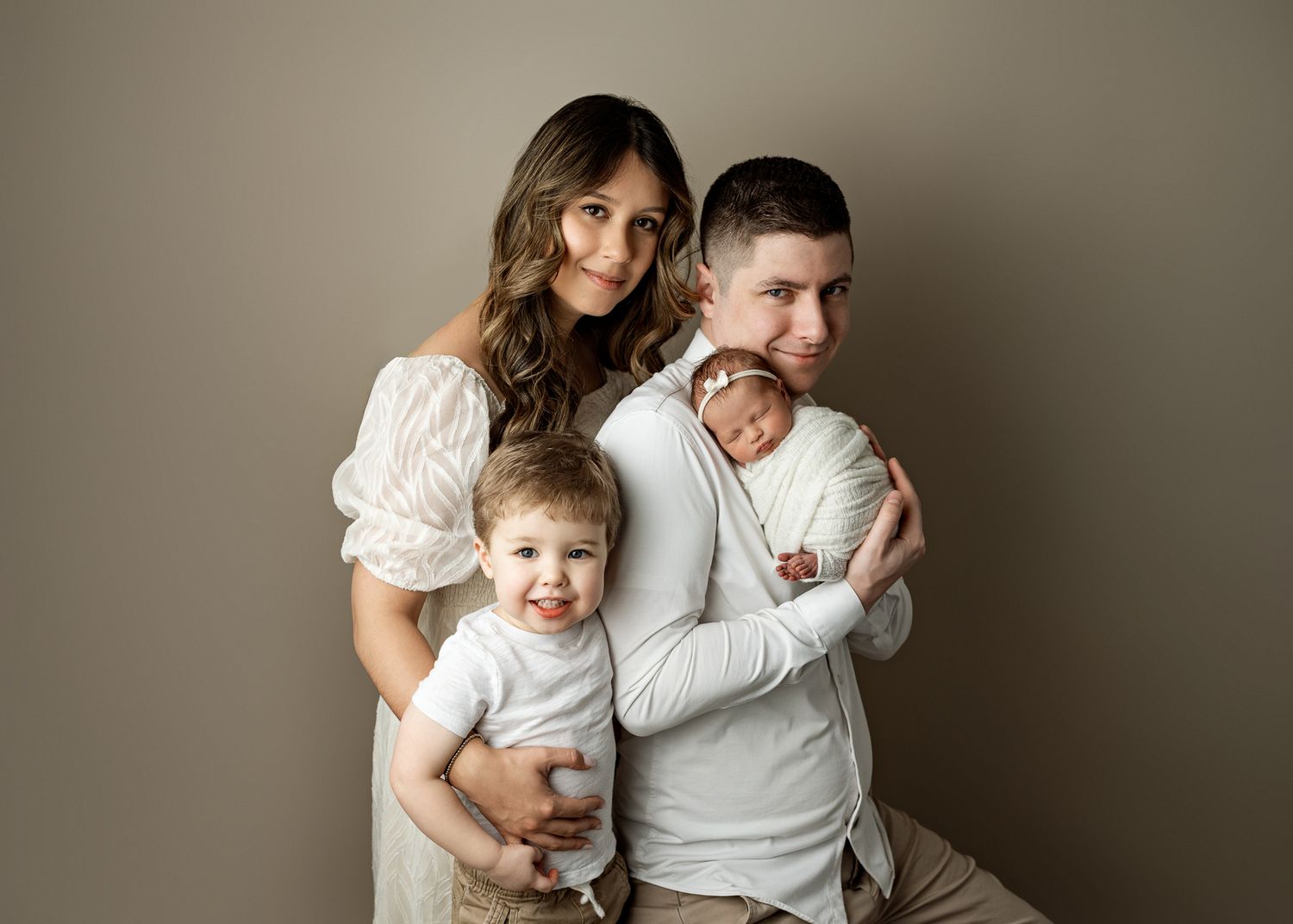 Family portrait in neutral tones with parents holding two young children against beige background.