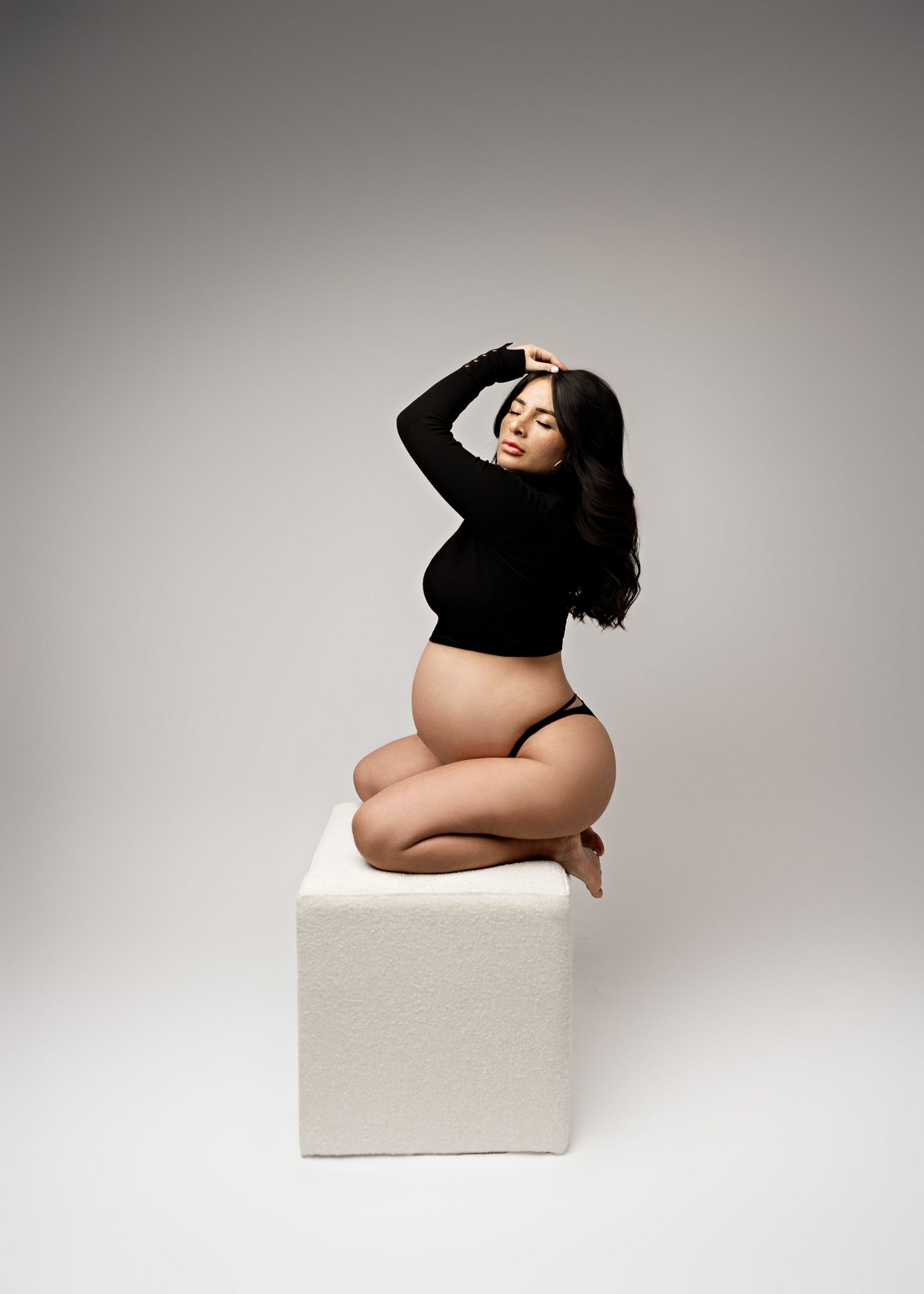 A maternity photo of someone in a black crop top posing gracefully on a white cube against a gray backdrop.