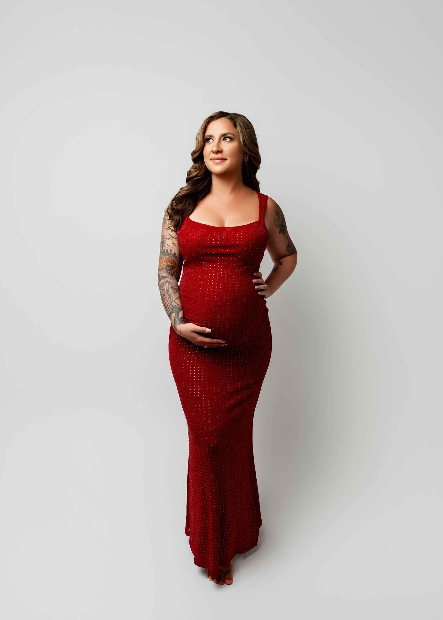 Pregnant woman in red maxi dress with tattoos posing for maternity photo on white background.