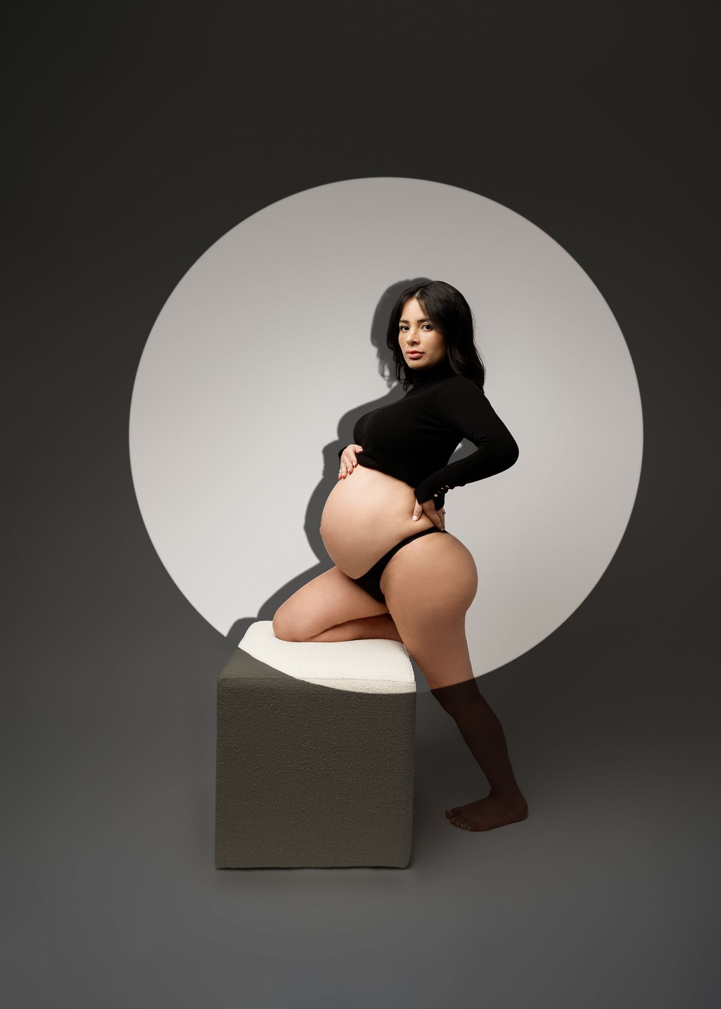 Dramatic maternity portrait with circular backdrop showing woman in black sweater on geometric platform.