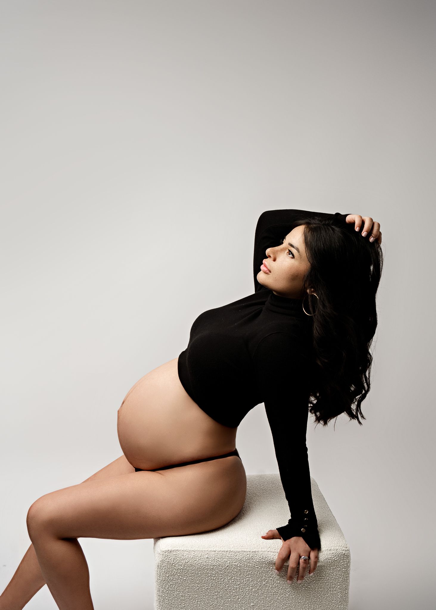 Artistic maternity photo of a woman in a black sweater posed on a white cube against light background.