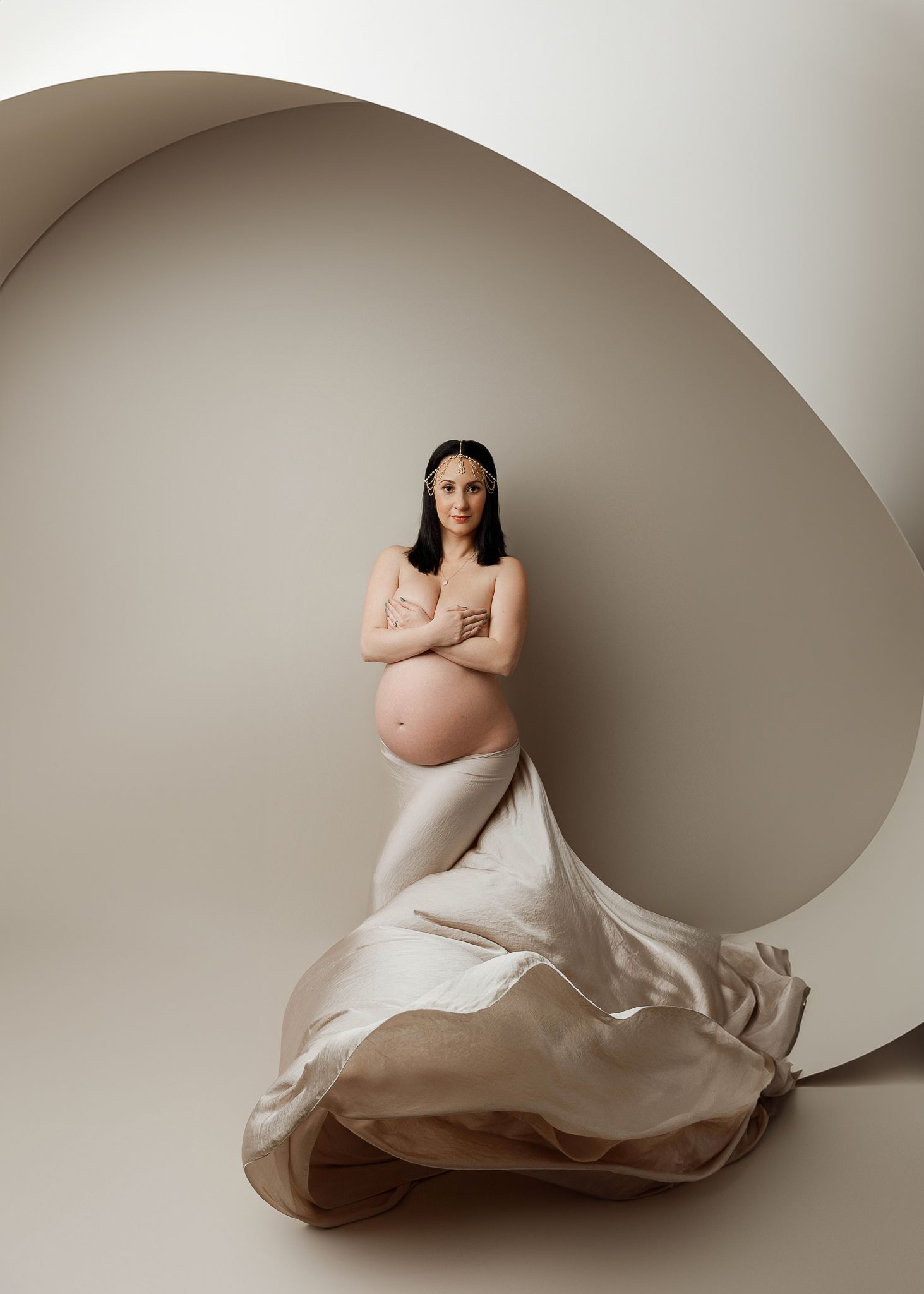 Artistic maternity photo of expectant mother in flowing white fabric against curved beige backdrop.