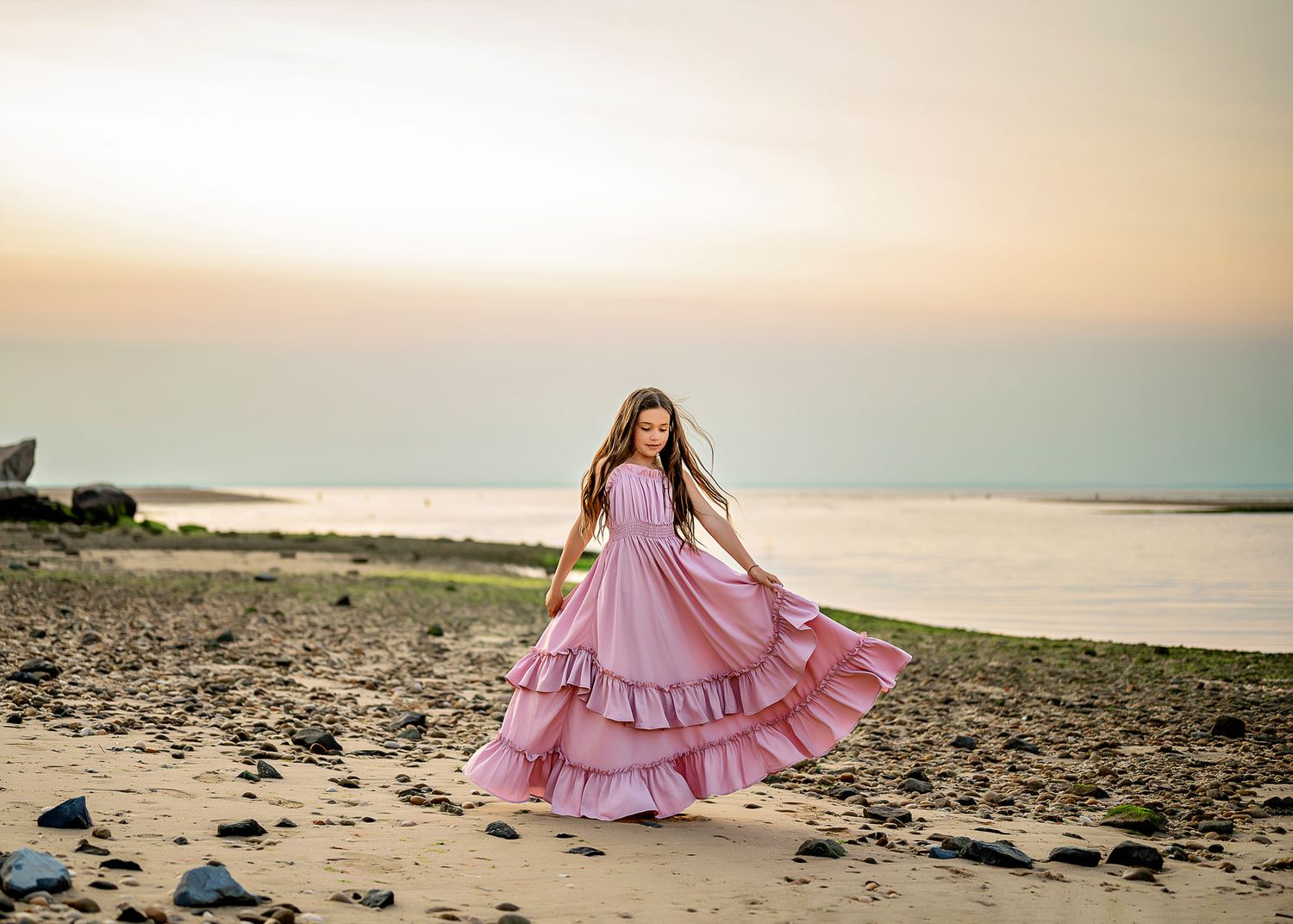 A person in a flowing pink tiered maxi dress twirls on a beach at sunset with ocean waves in the background.