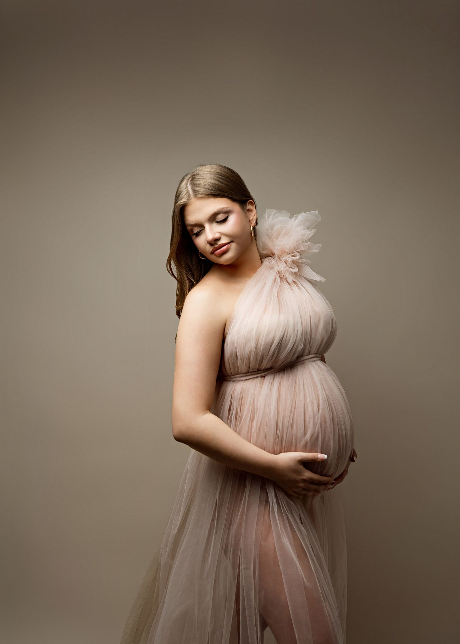 Elegant maternity photo series showing a woman in a flowing blush tulle gown against a neutral studio backdrop.