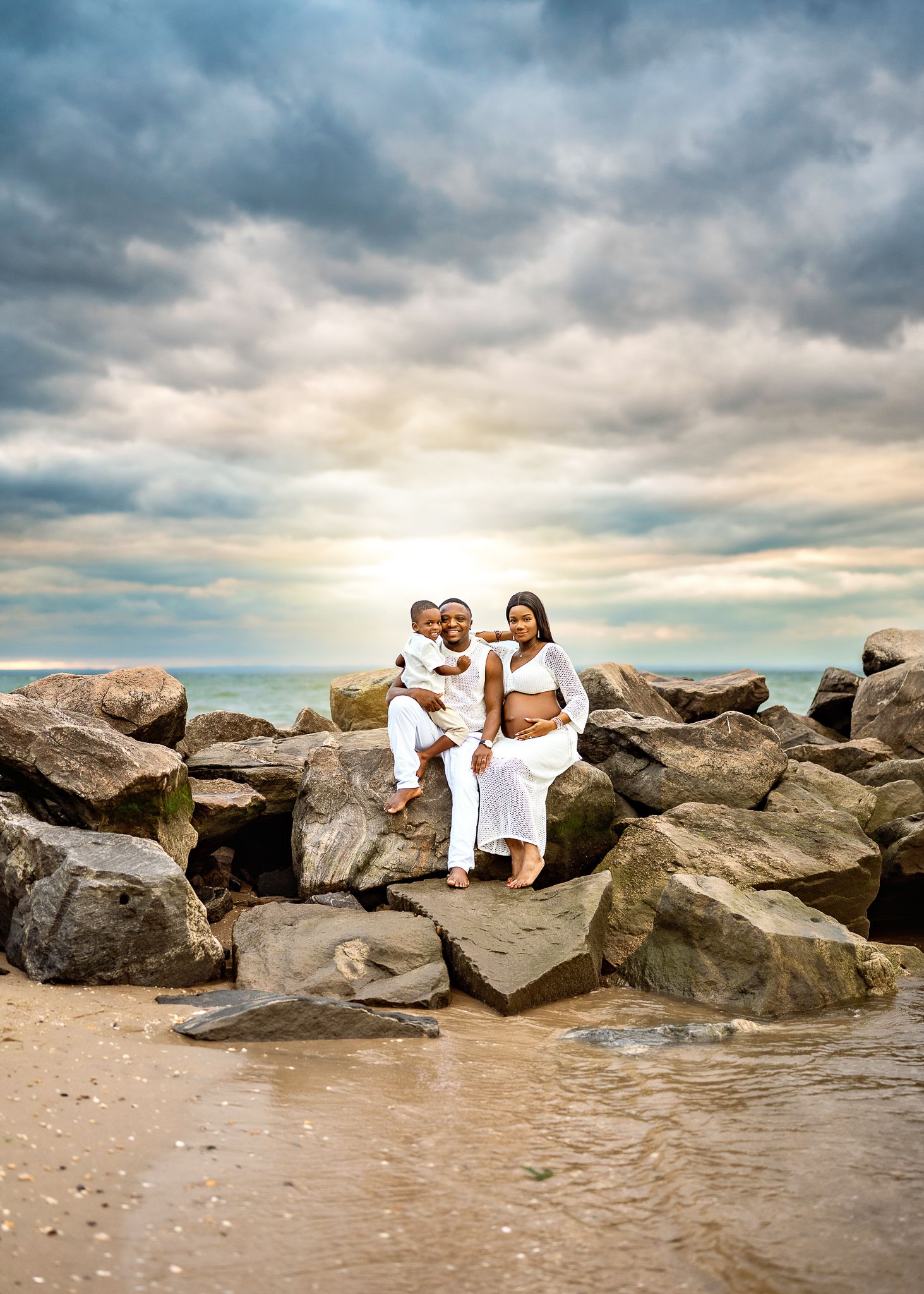 People sit together on rocky beach at sunset with dramatic cloudy sky and ocean waves in background.
