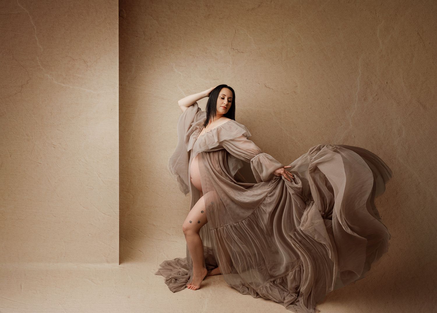 Maternity photo featuring flowy taupe dress billowing dramatically against neutral backdrop.