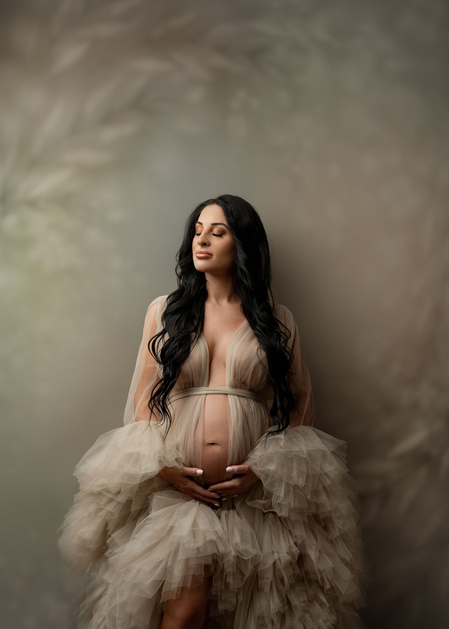 Beautiful maternity portrait in flowing cream tulle gown against moody backdrop shows expecting mother cradling belly.