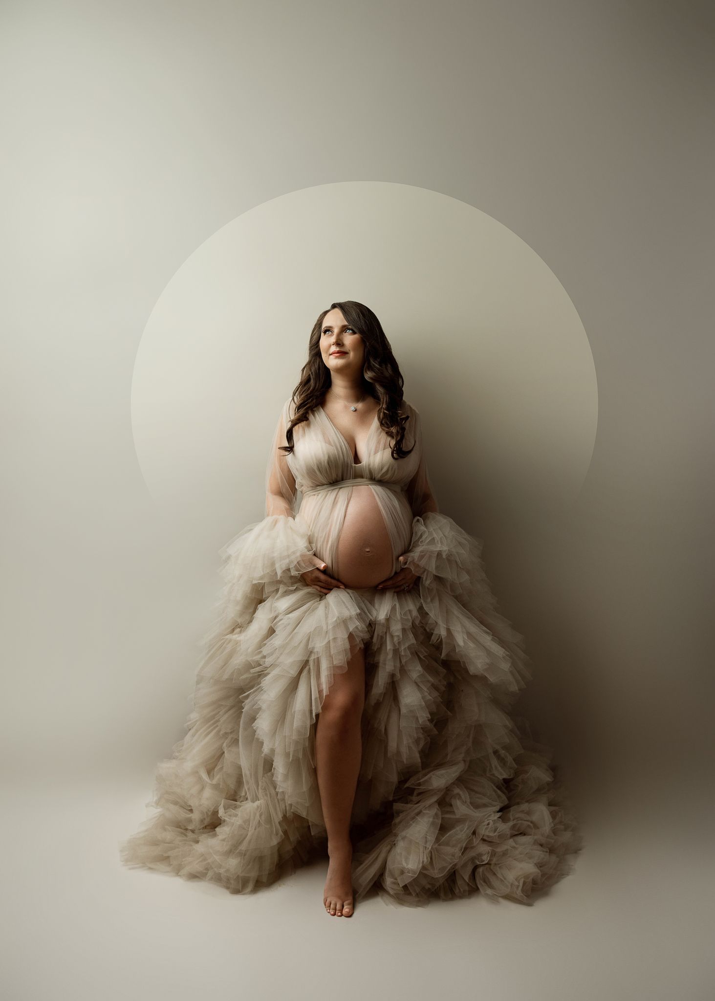 Artistic maternity photo of an expectant mother in a flowing tulle gown posing against a minimalist circular backdrop.