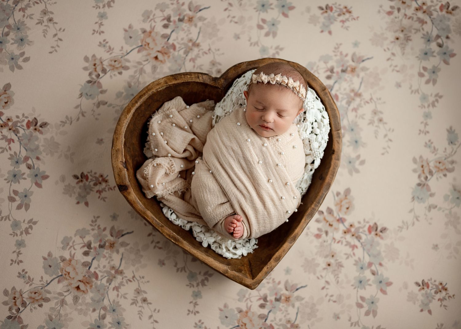 A sleeping newborn baby wearing white lace clothing lies in a rustic heart-shaped wooden bowl against floral wallpaper.