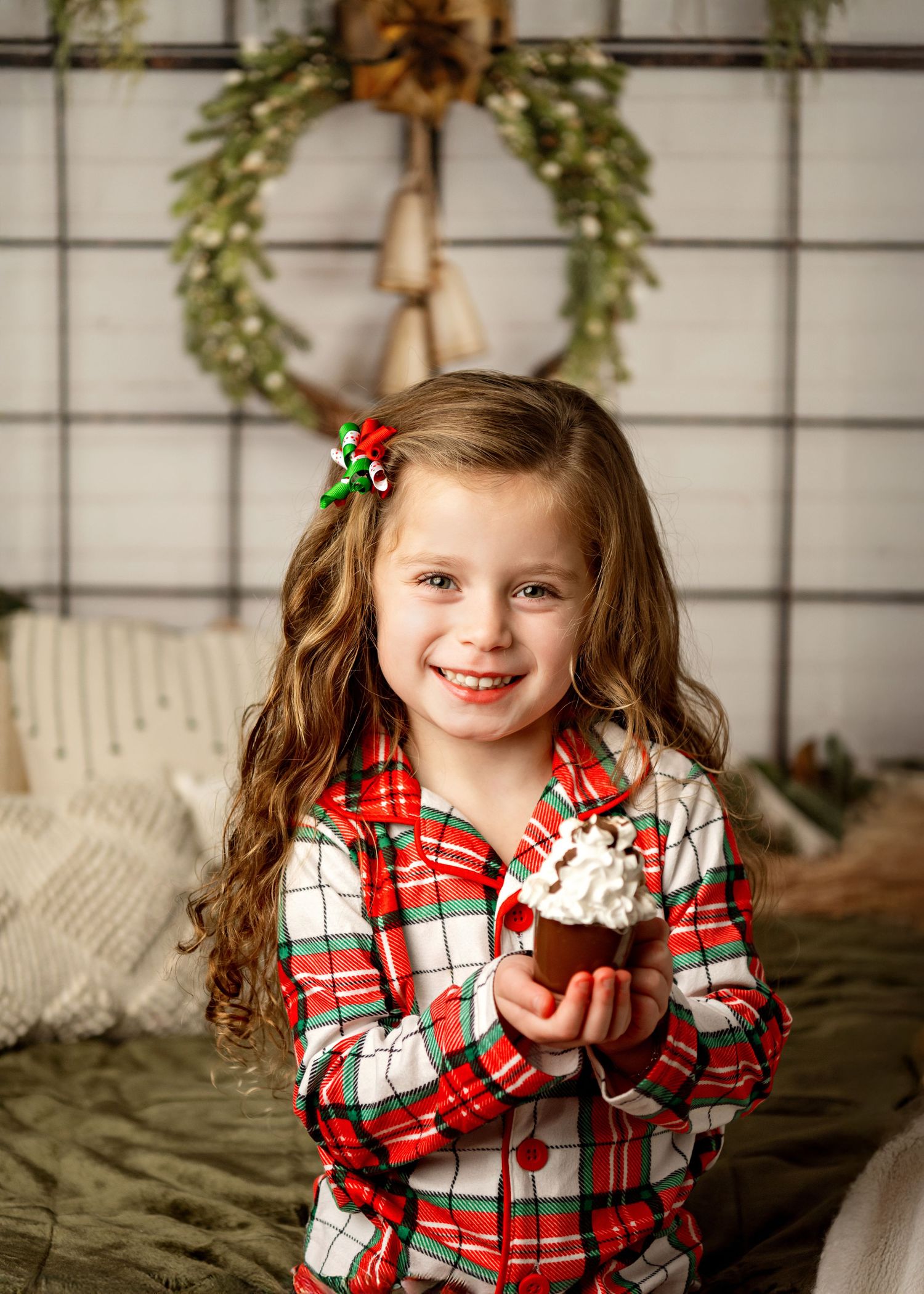 Child wearing plaid red and white holiday flannel shirt holds cupcake in front of decorative winter wreath backdrop.