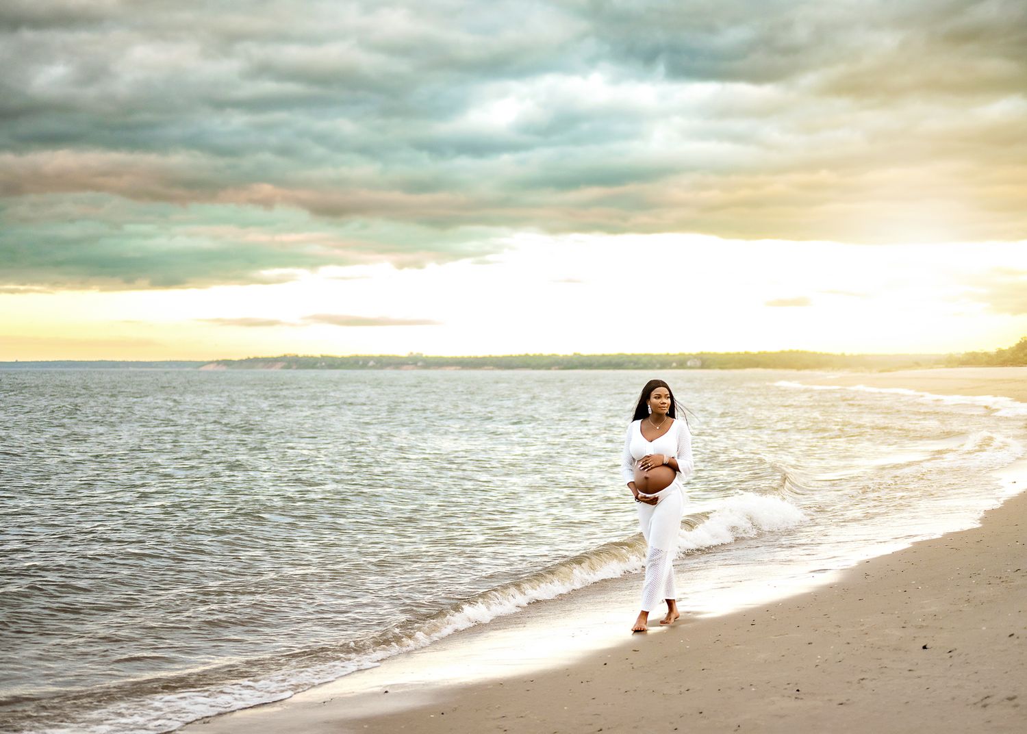 A person in white clothing walks along a serene beach at sunset with dramatic cloudy skies overhead.